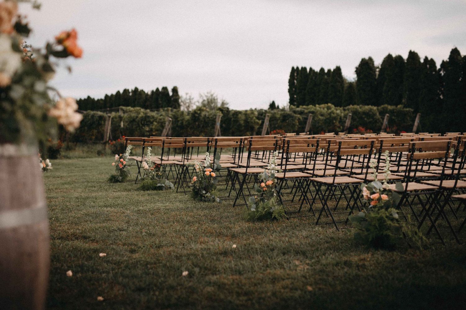Outdoor wedding ceremony setup with rows of black chairs on green lawn beside vineyard.