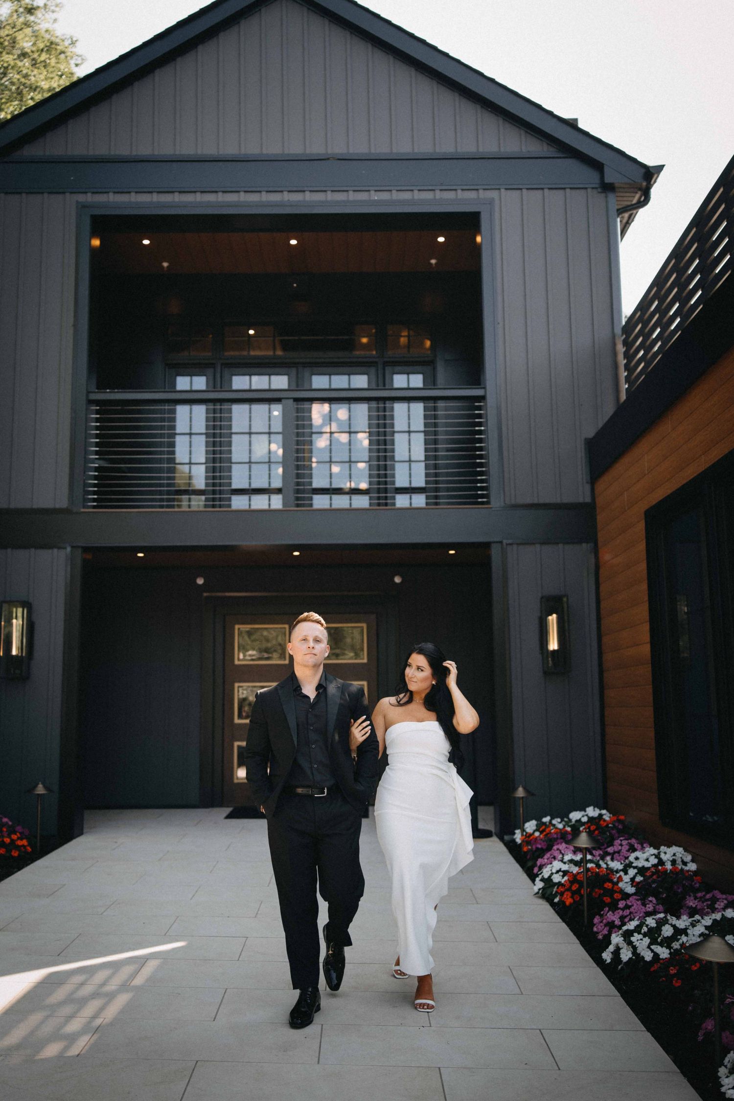 Stylish couple walking in front of modern dark gray barn-style building, woman in white dress.