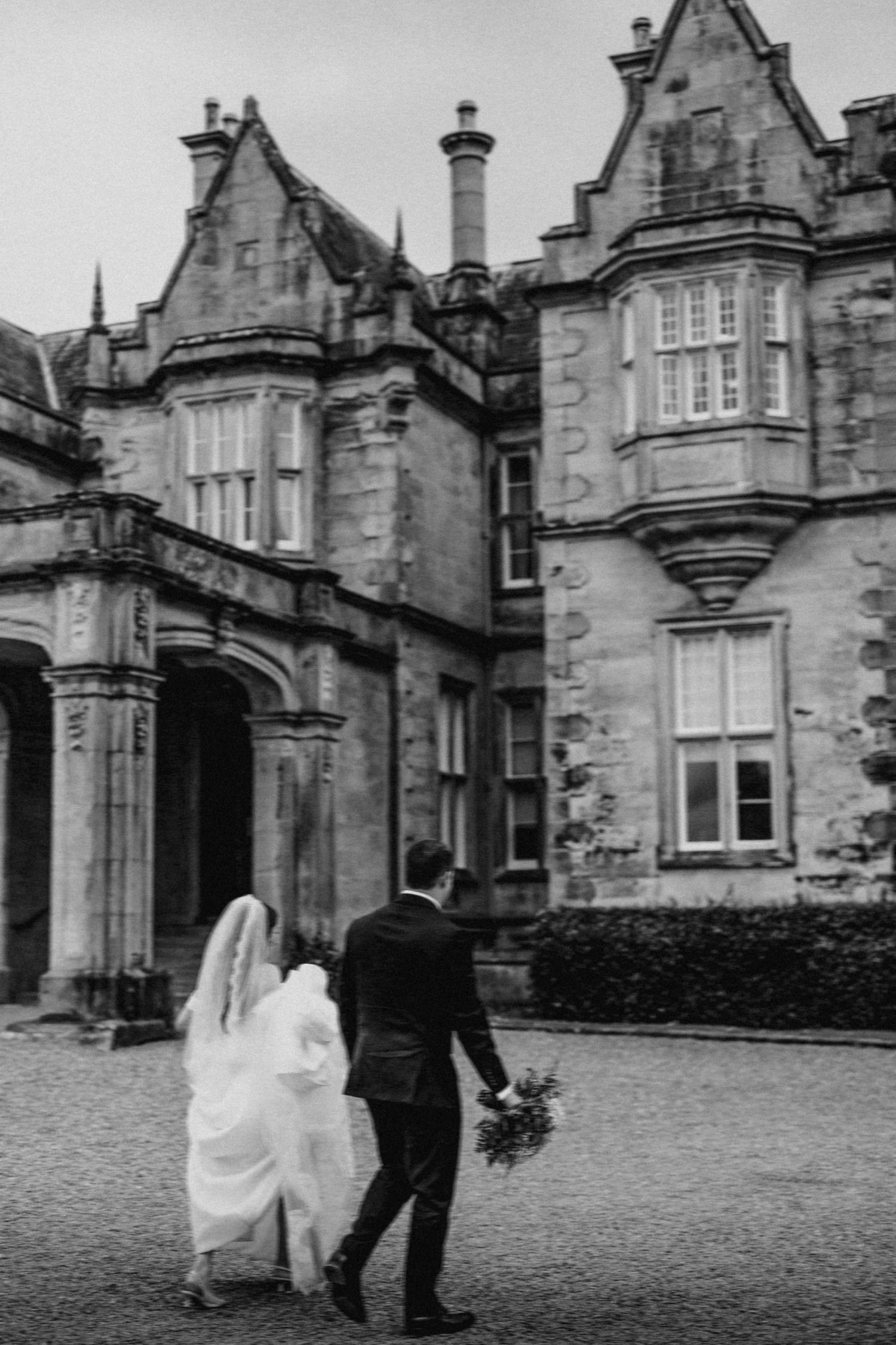 Bride and groom walking hand-in-hand toward a grand historic castle venue in a black and white wedding photo.