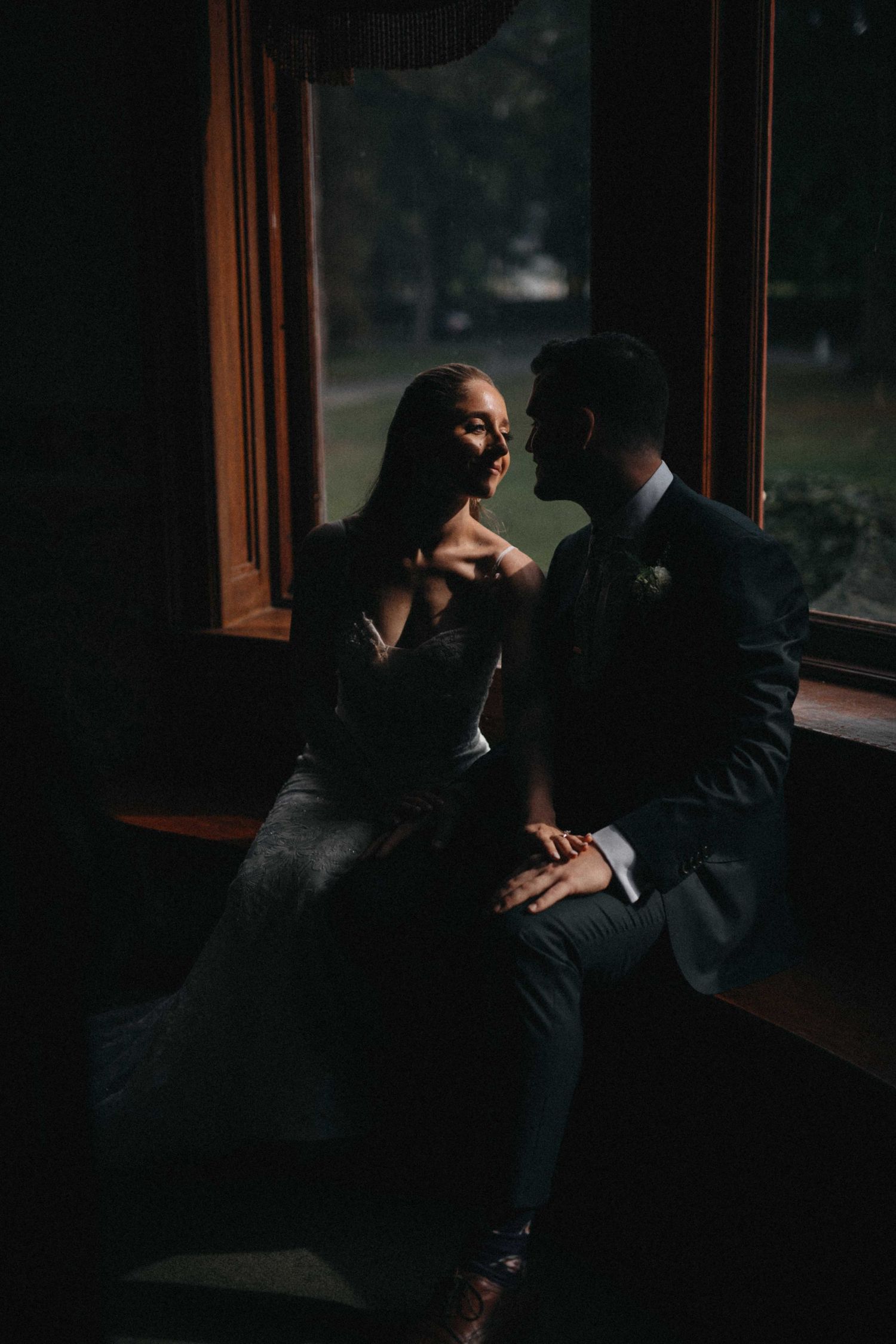 Bride and groom share intimate moment seated by wooden window in dramatic moody wedding portrait.
