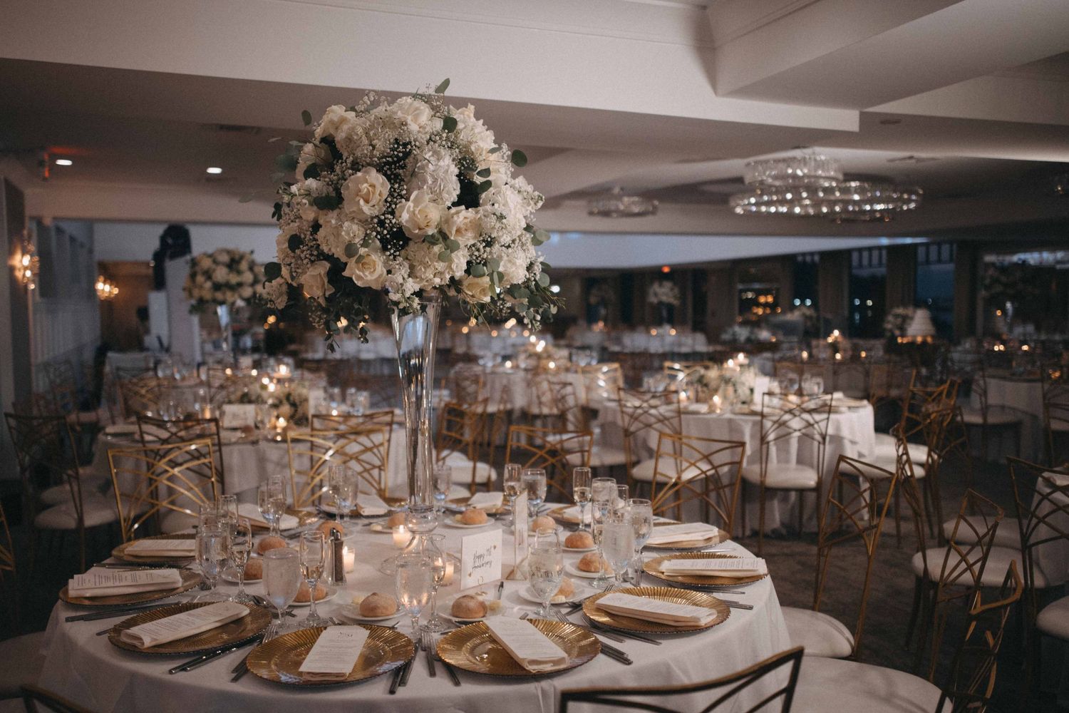 Elegant wedding reception with tall white floral centerpieces, gold chargers, and crystal chandeliers.