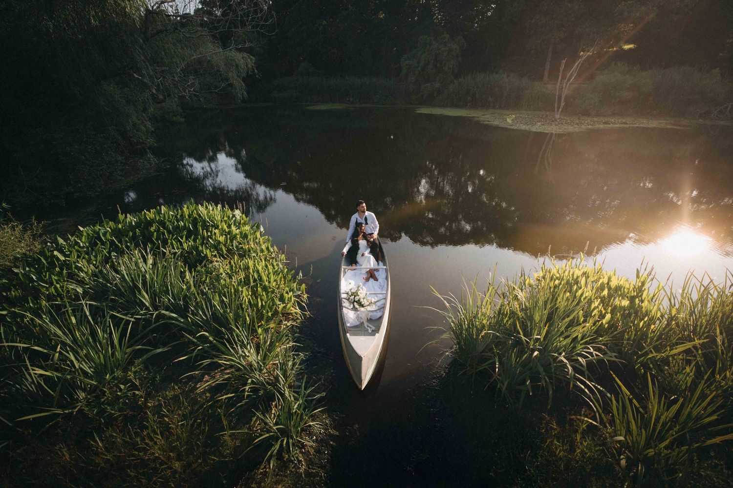 Aerial view of a couple in a white canoe on a calm, lush green waterway at golden hour.