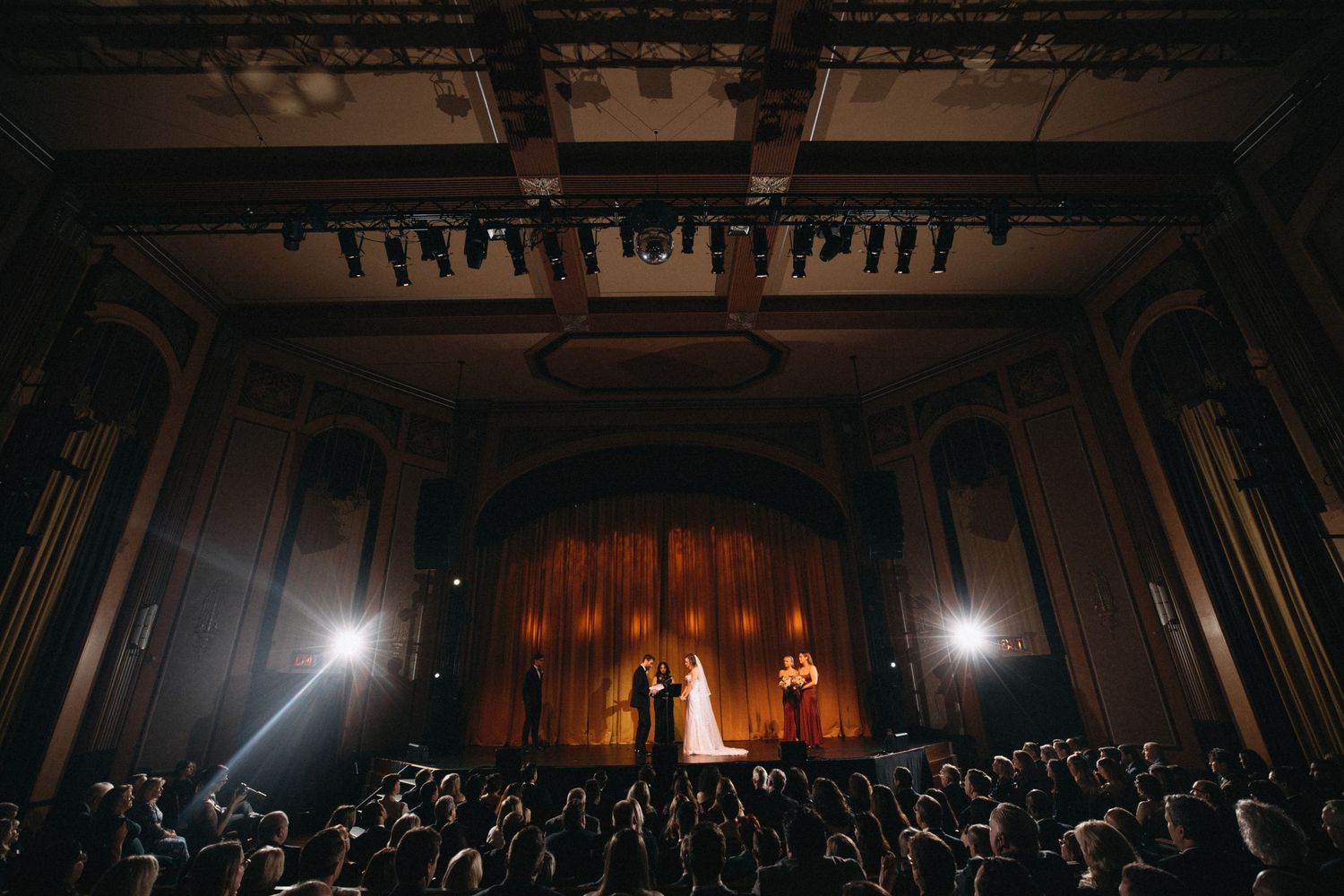 Wide shot of bride and groom exchanging vows on stage in stunning historic theater ceremony.