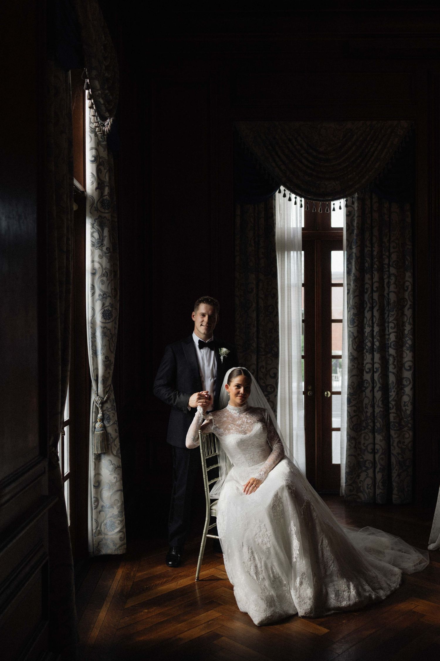 Elegant bride seated in chair with groom standing beside her near tall windows in dark interior.