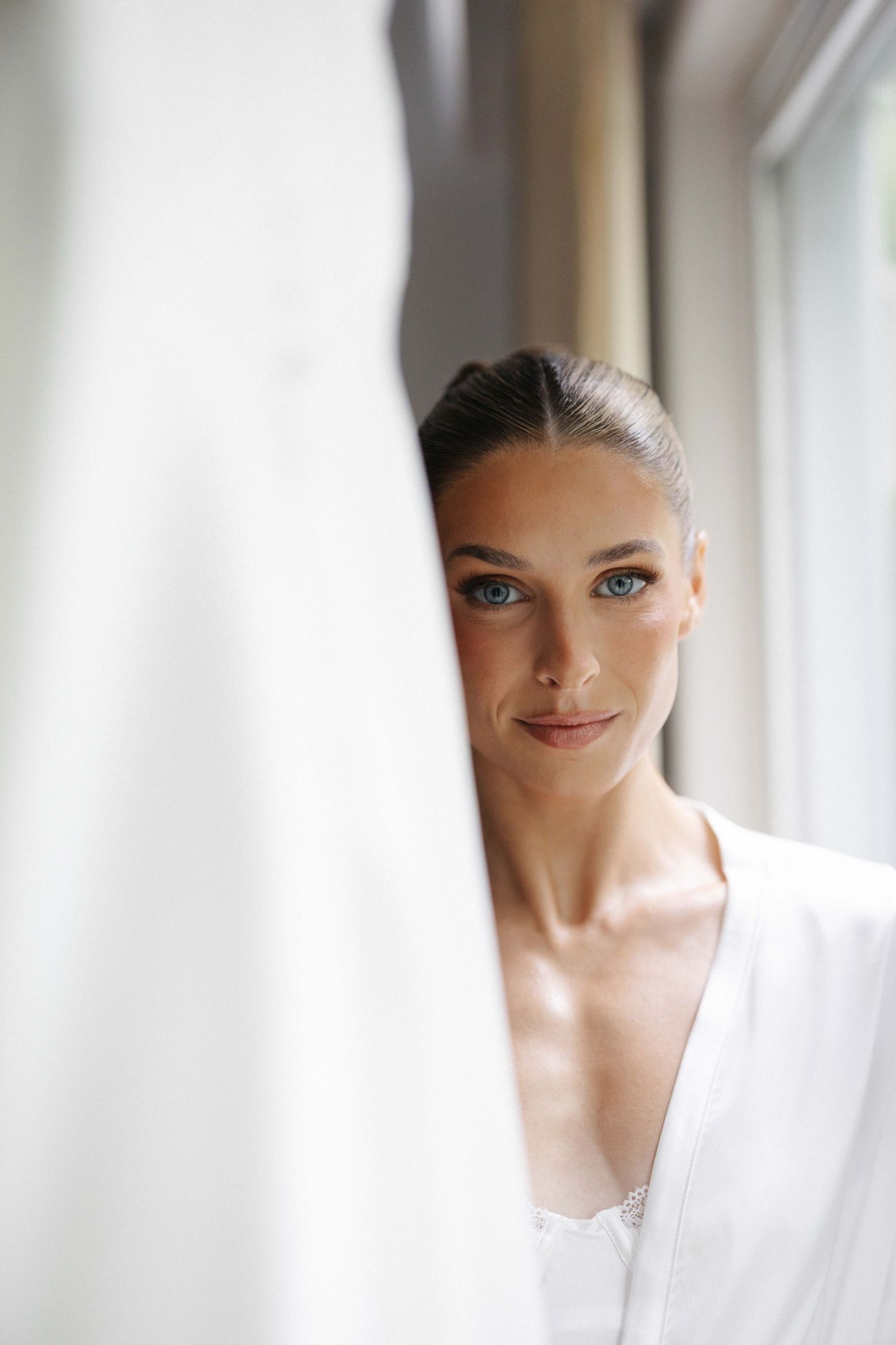 A bride in a white robe peers elegantly from behind a wedding dress near a sunlit window.
