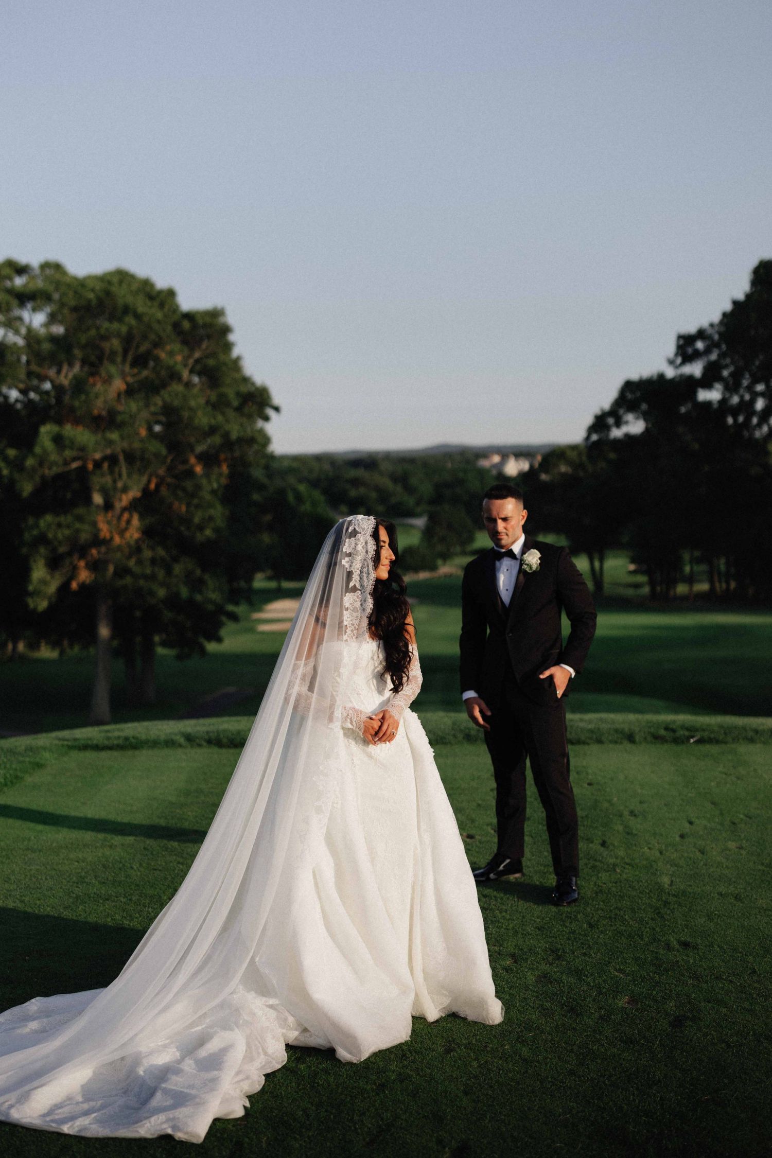 Bride in white gown with long veil stands beside groom in black tuxedo on lush green golf course at sunset.