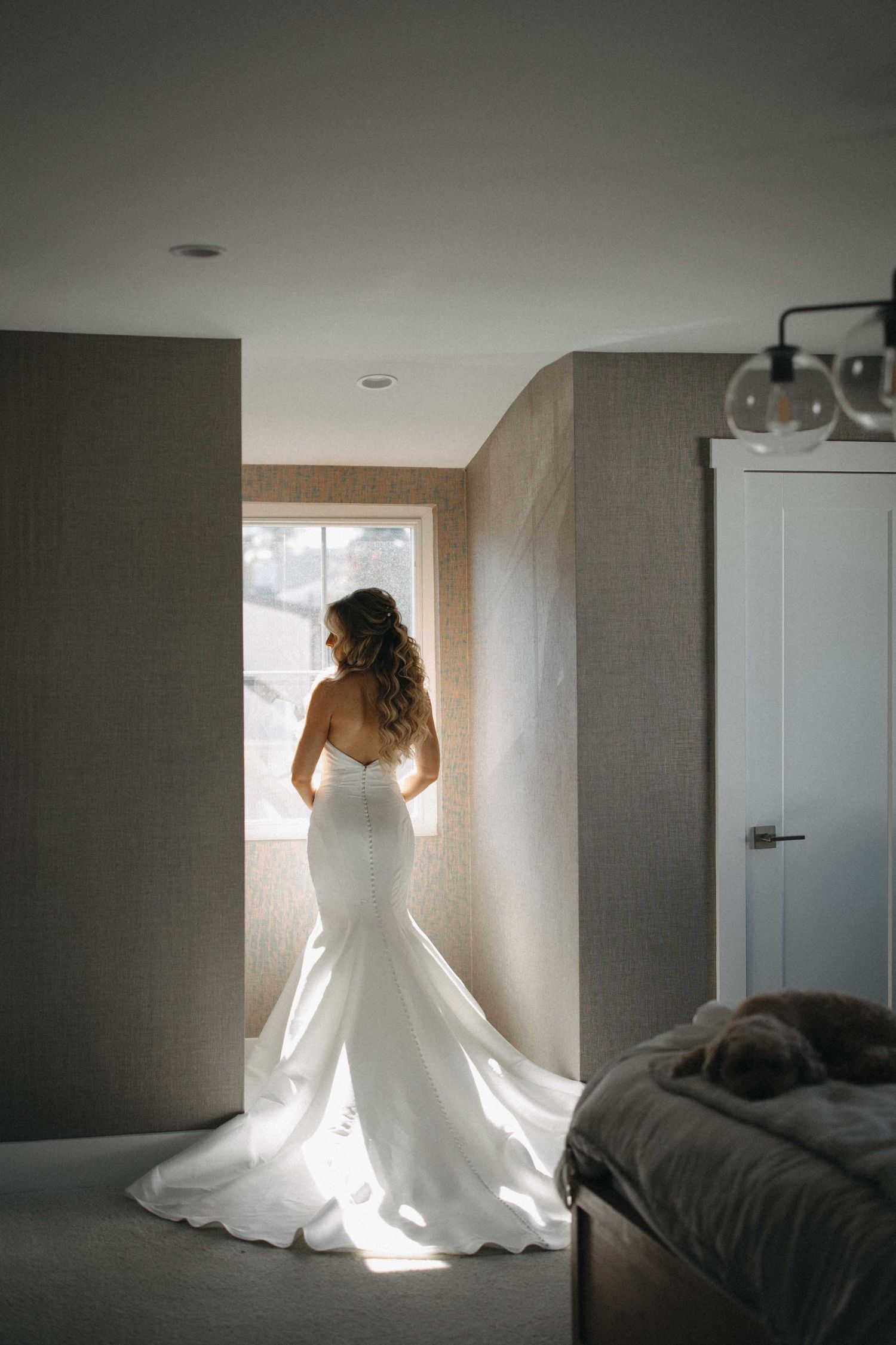 Bride in strapless mermaid wedding gown with train standing by window in soft backlight.