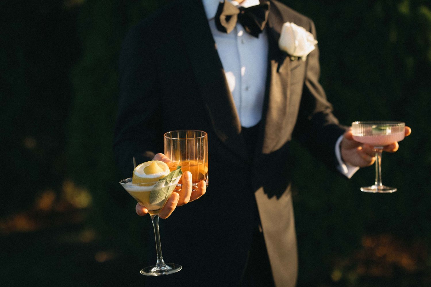 Elegantly dressed wedding guest holding champagne flutes and cocktail glasses at golden hour.