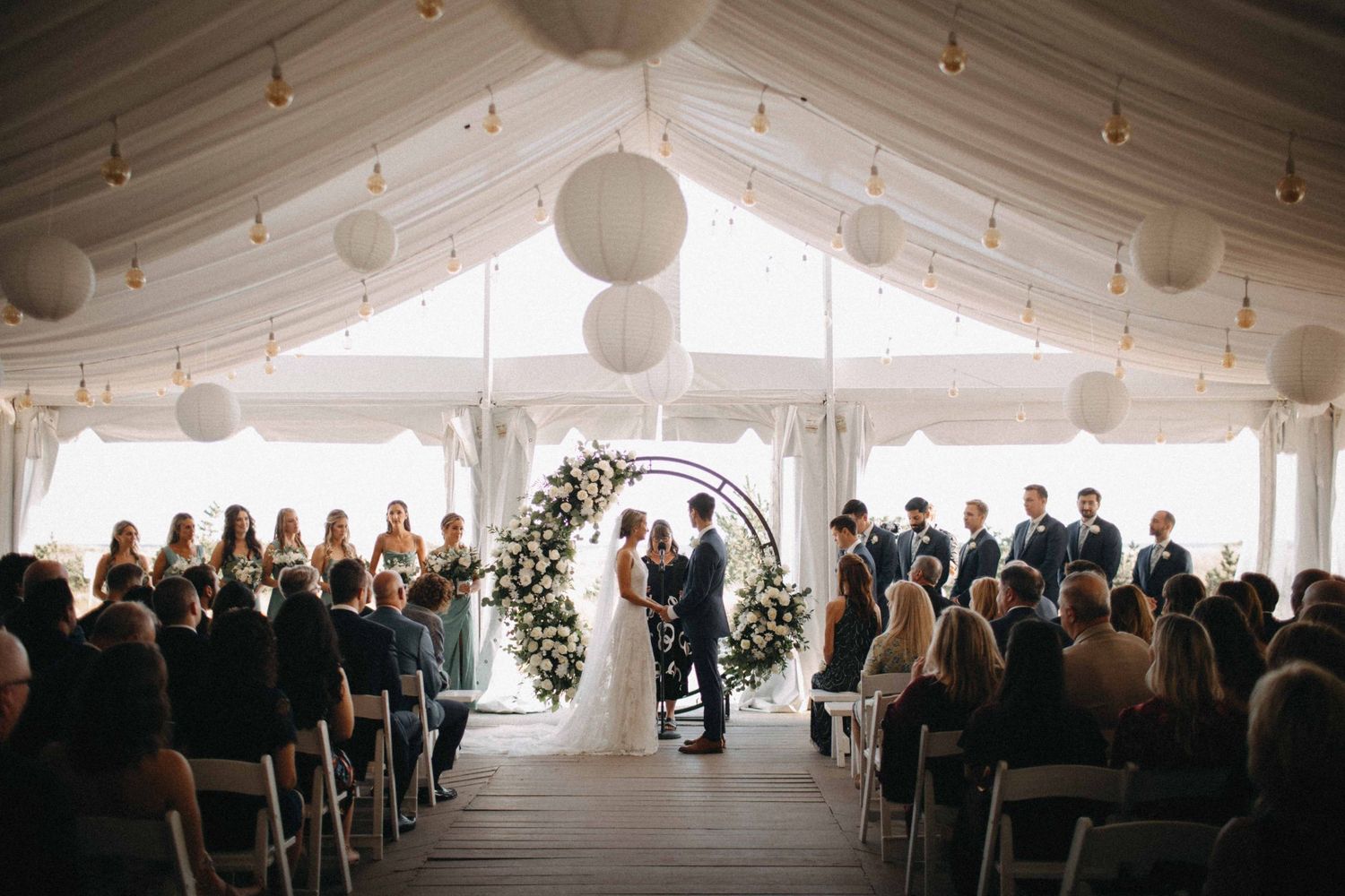 Bride and groom exchange vows under floral arch in elegant tented oceanfront wedding ceremony.