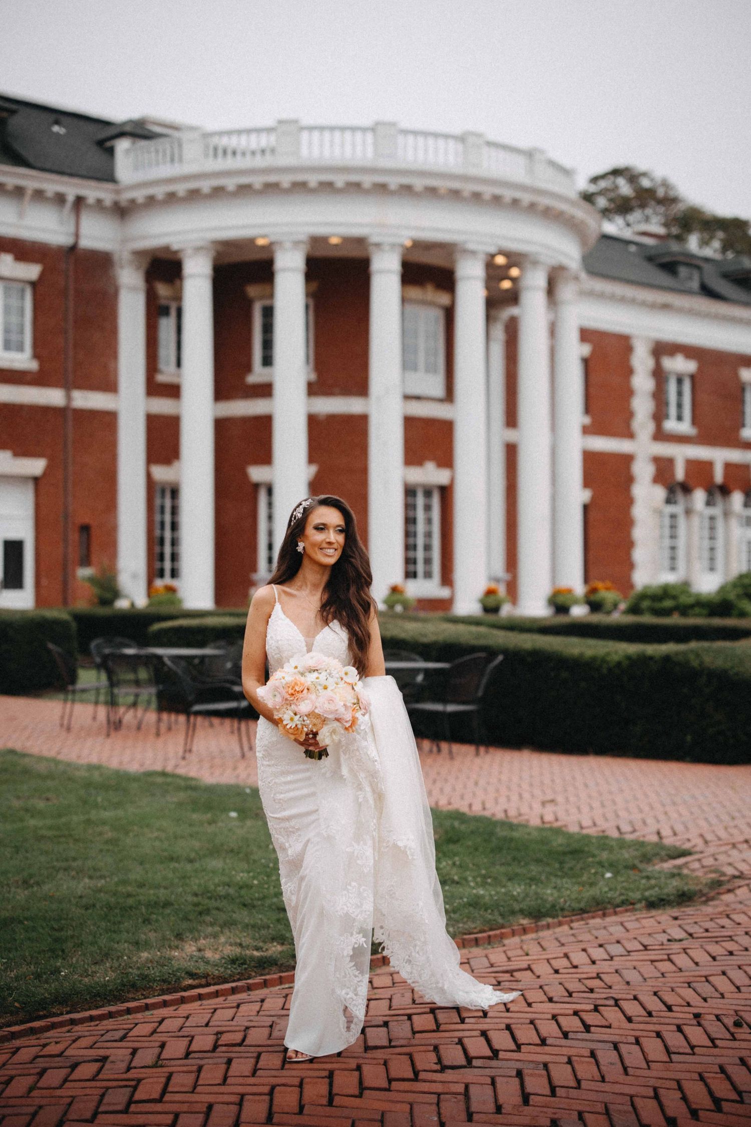 Bride in lace gown holds bouquet in front of a brick colonial mansion with white columns.