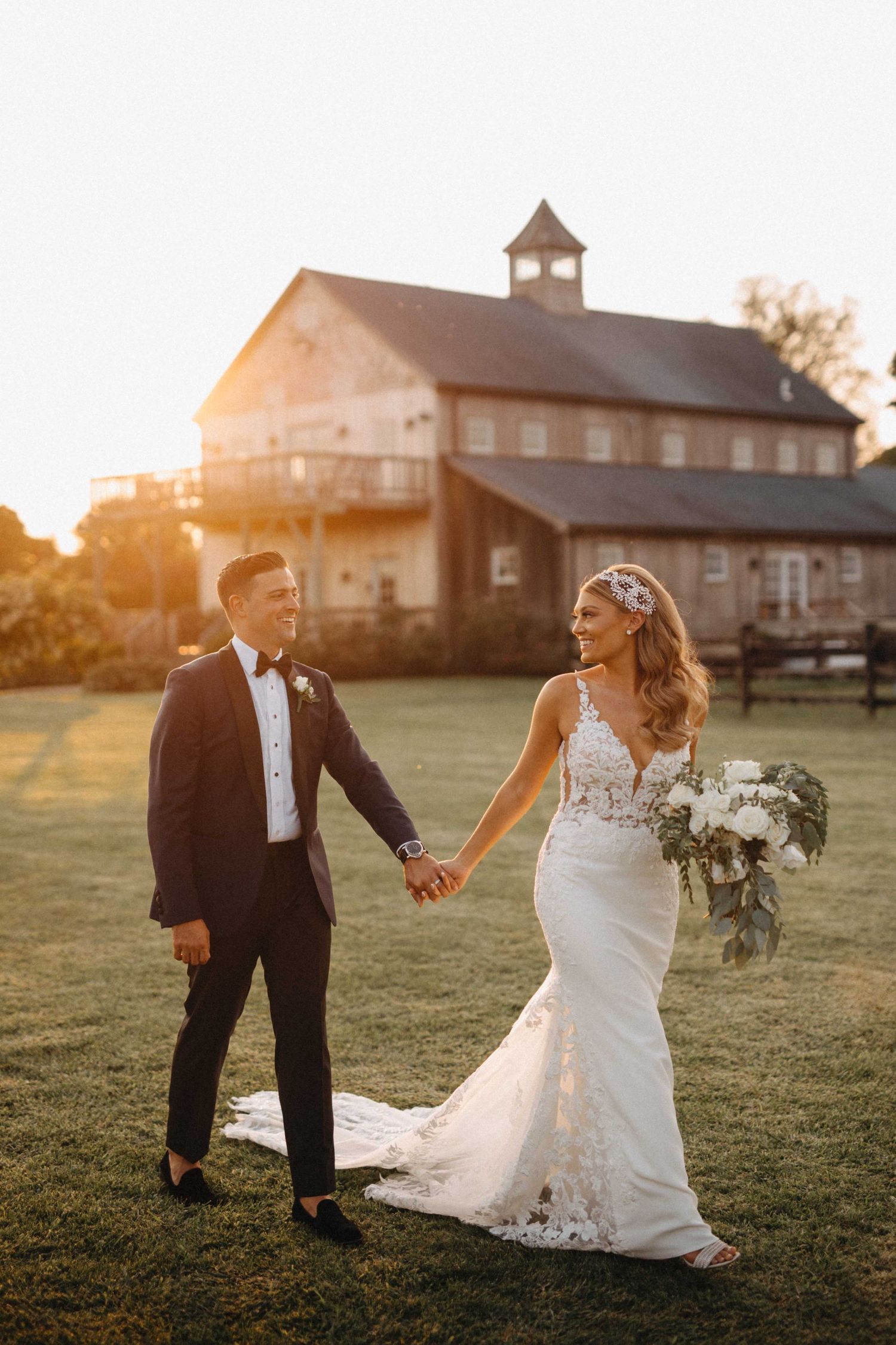 Bride and groom holding hands at golden sunset with rustic barn venue in background.