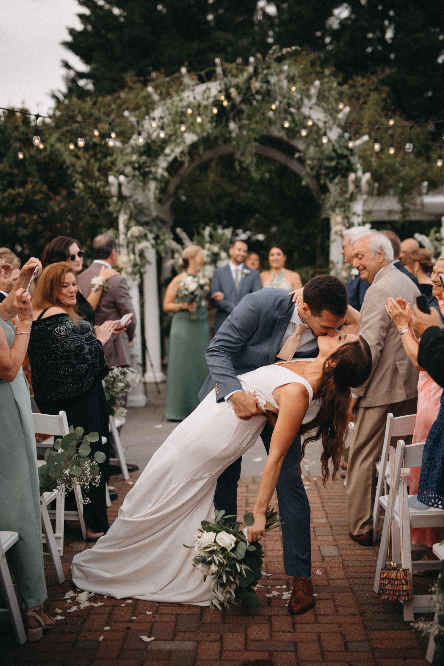 Bride and groom share romantic dip kiss while walking down brick aisle as guests celebrate outdoor wedding ceremony.