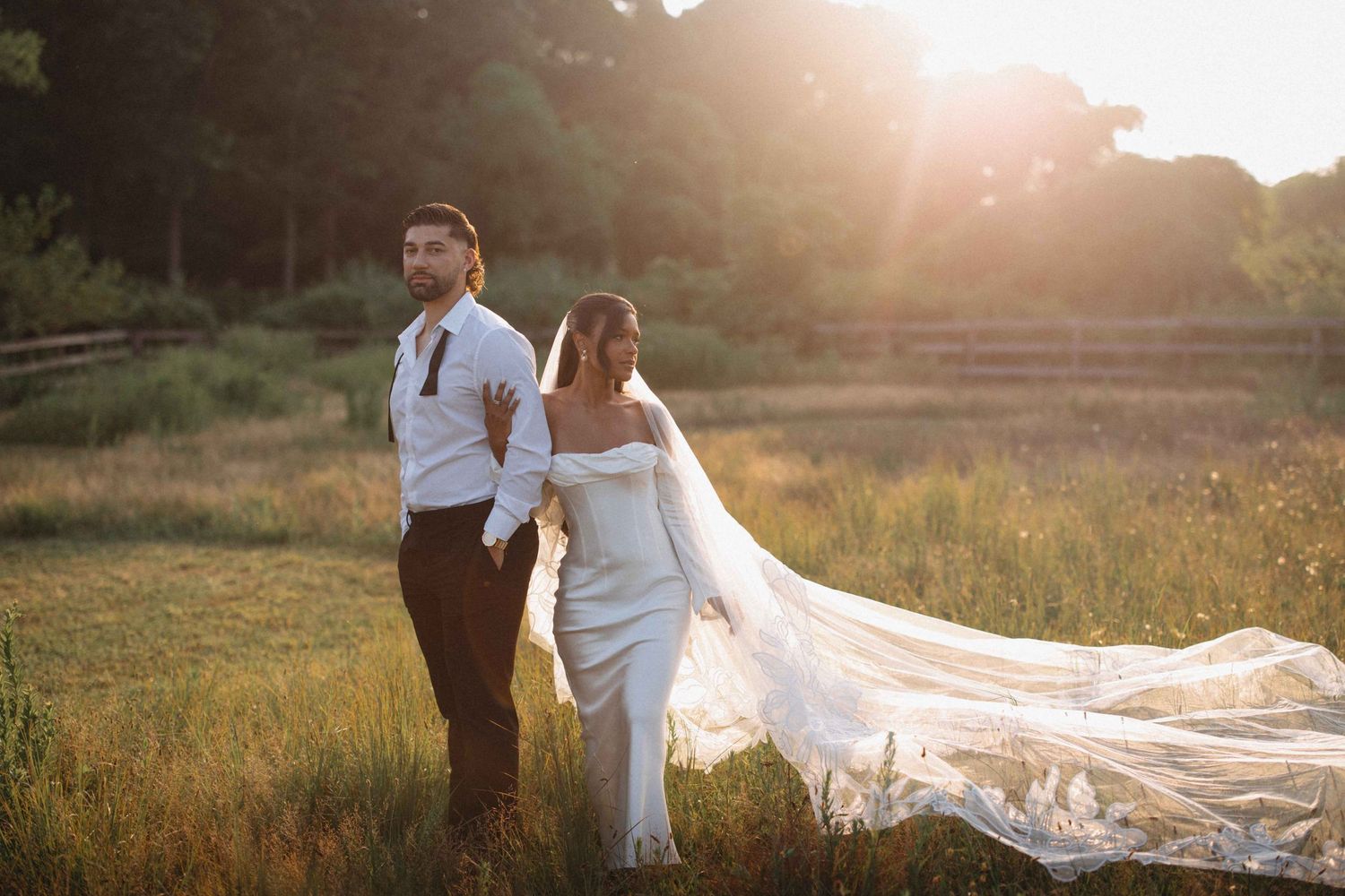 Bride in a flowing off-shoulder gown with a long train standing with groom at golden sunset.