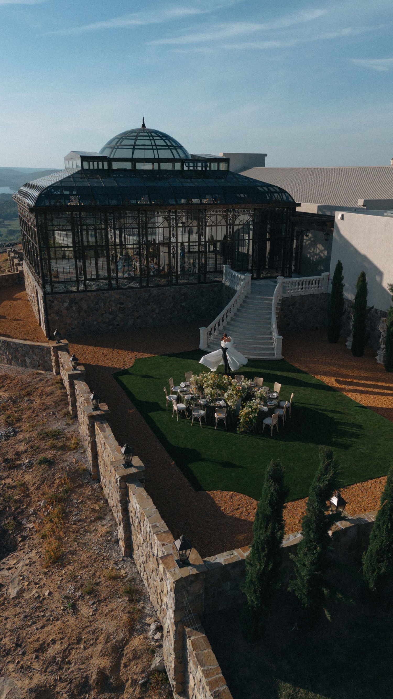 Aerial view of an elegant glass greenhouse wedding venue on a hilltop with outdoor reception tables and manicured gardens.