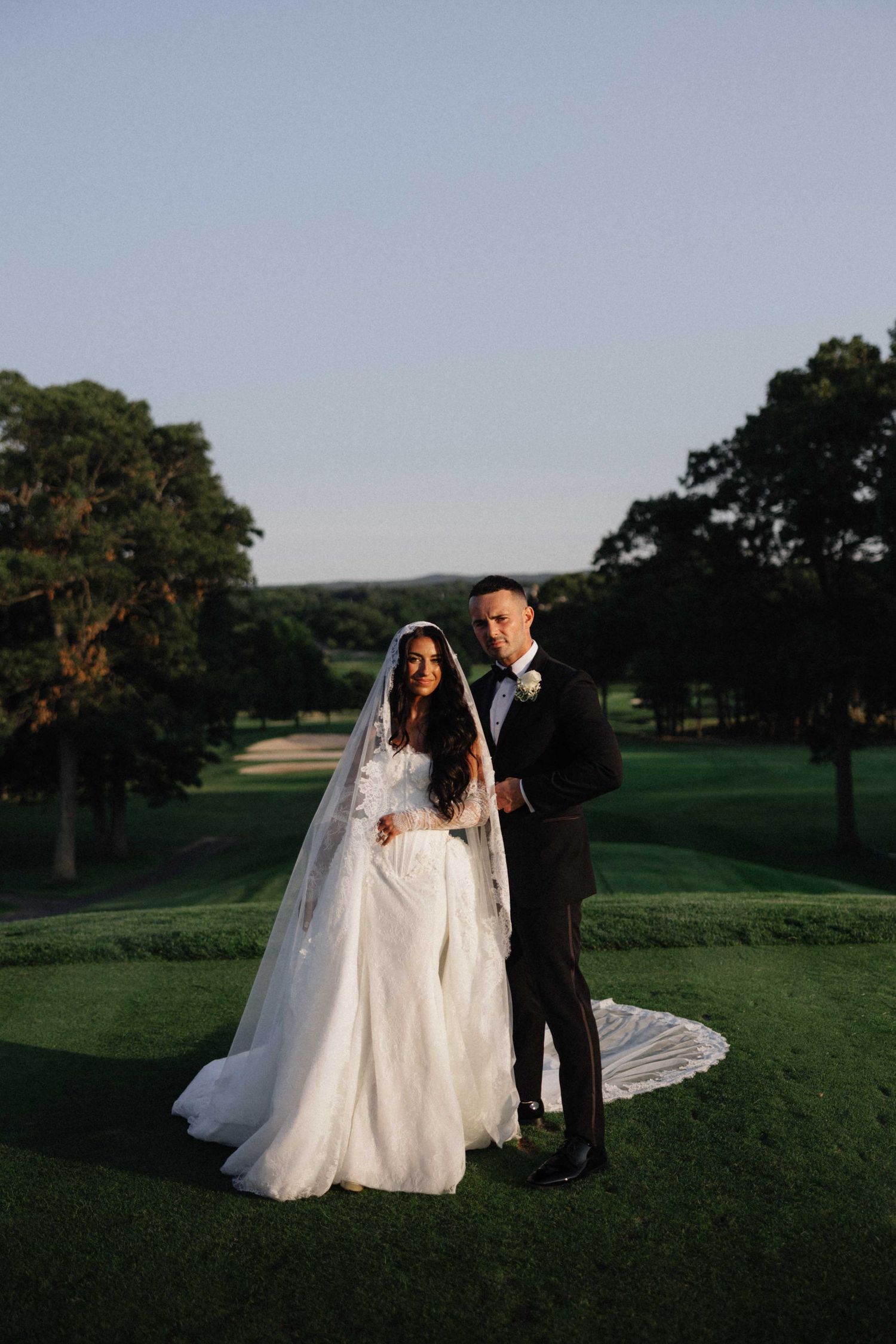 Bride in white lace gown with veil and groom in black tuxedo posing on lush green golf course at golden hour.