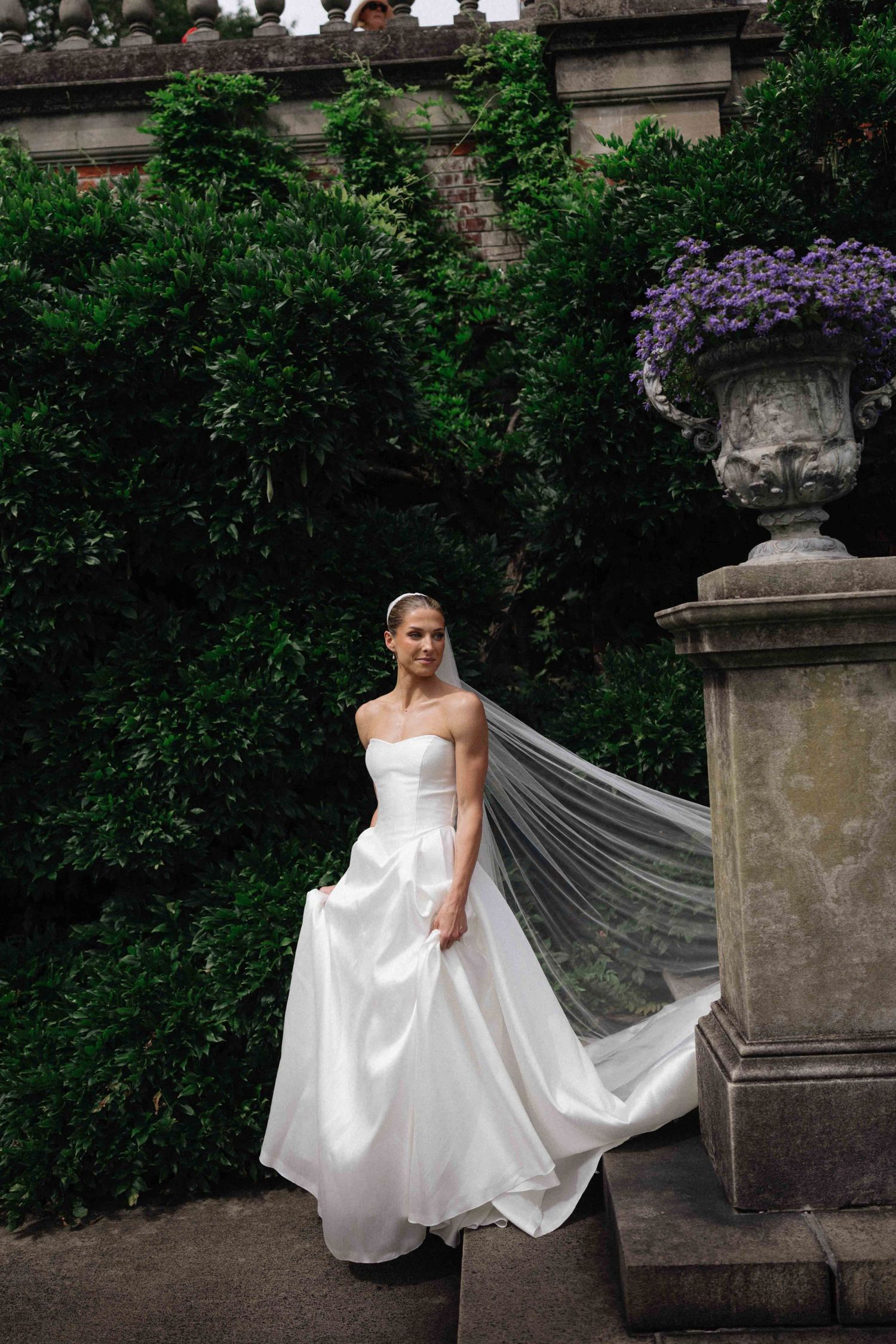 Bride in strapless white ball gown with flowing veil stands on stone steps surrounded by lush greenery and purple flowers.
