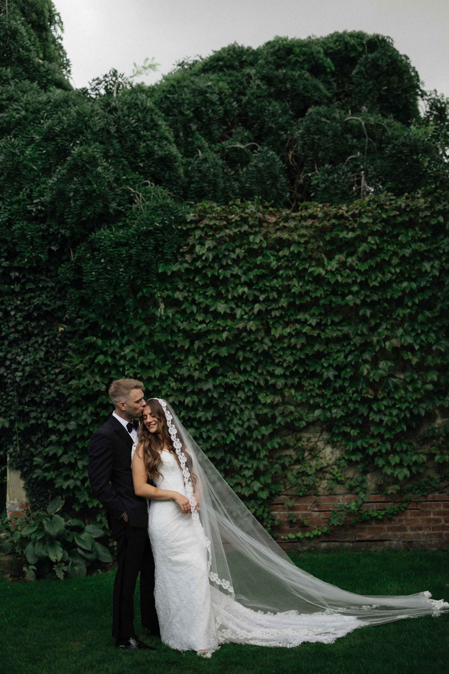 Bride and groom embrace in front of ivy-covered brick wall, bride wearing lace gown with cathedral veil.