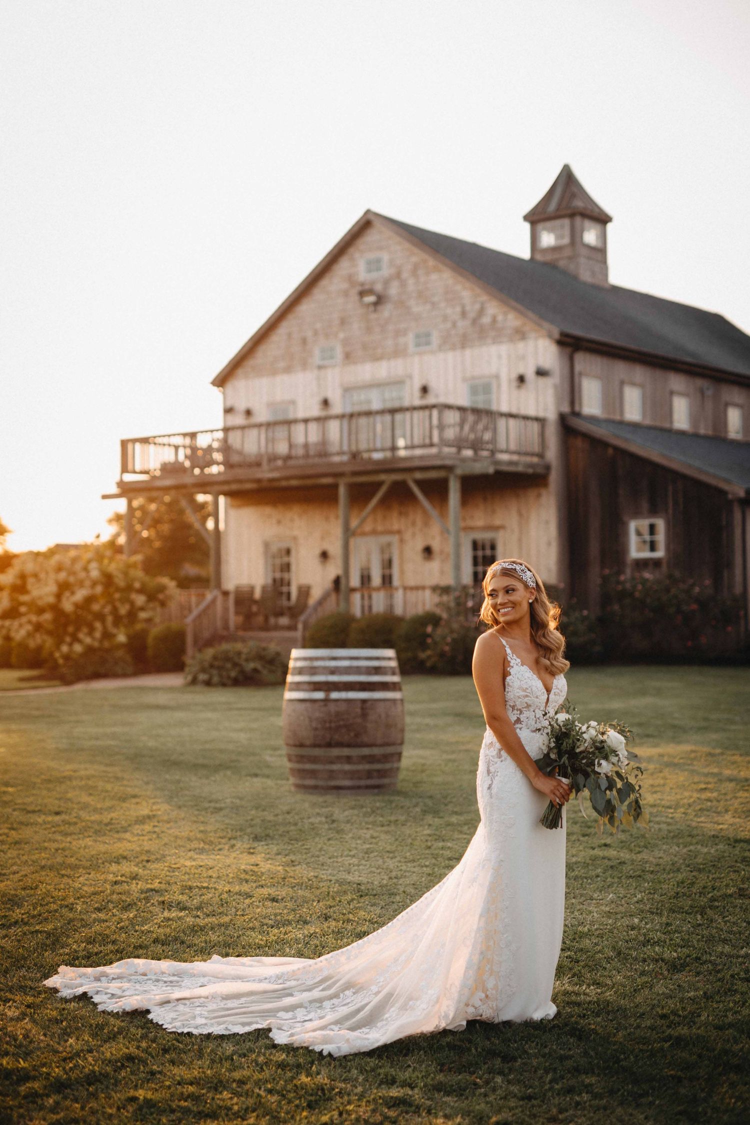 Bride in white lace gown holding bouquet stands before rustic barn venue at golden hour.
