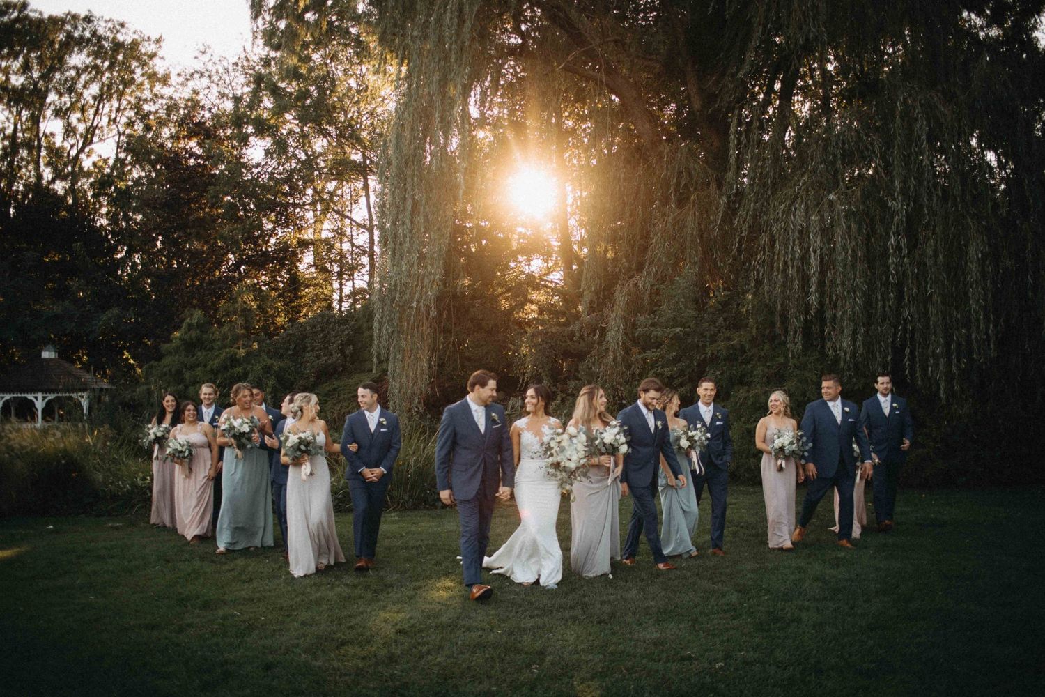 Wedding party walks together across green lawn at golden hour with large trees and white pergola in background.
