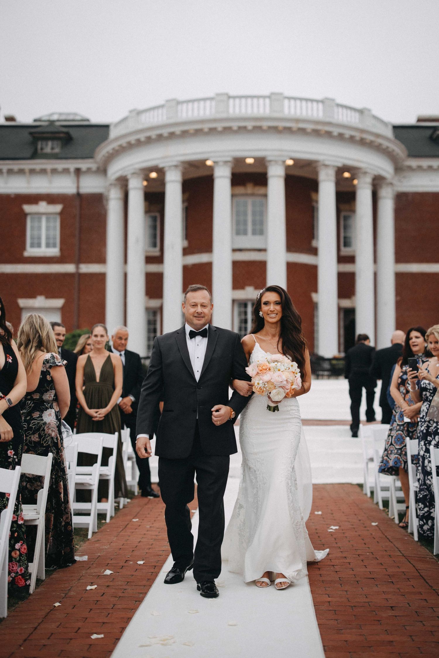 Father escorts bride down the aisle at an outdoor wedding ceremony at a brick mansion.