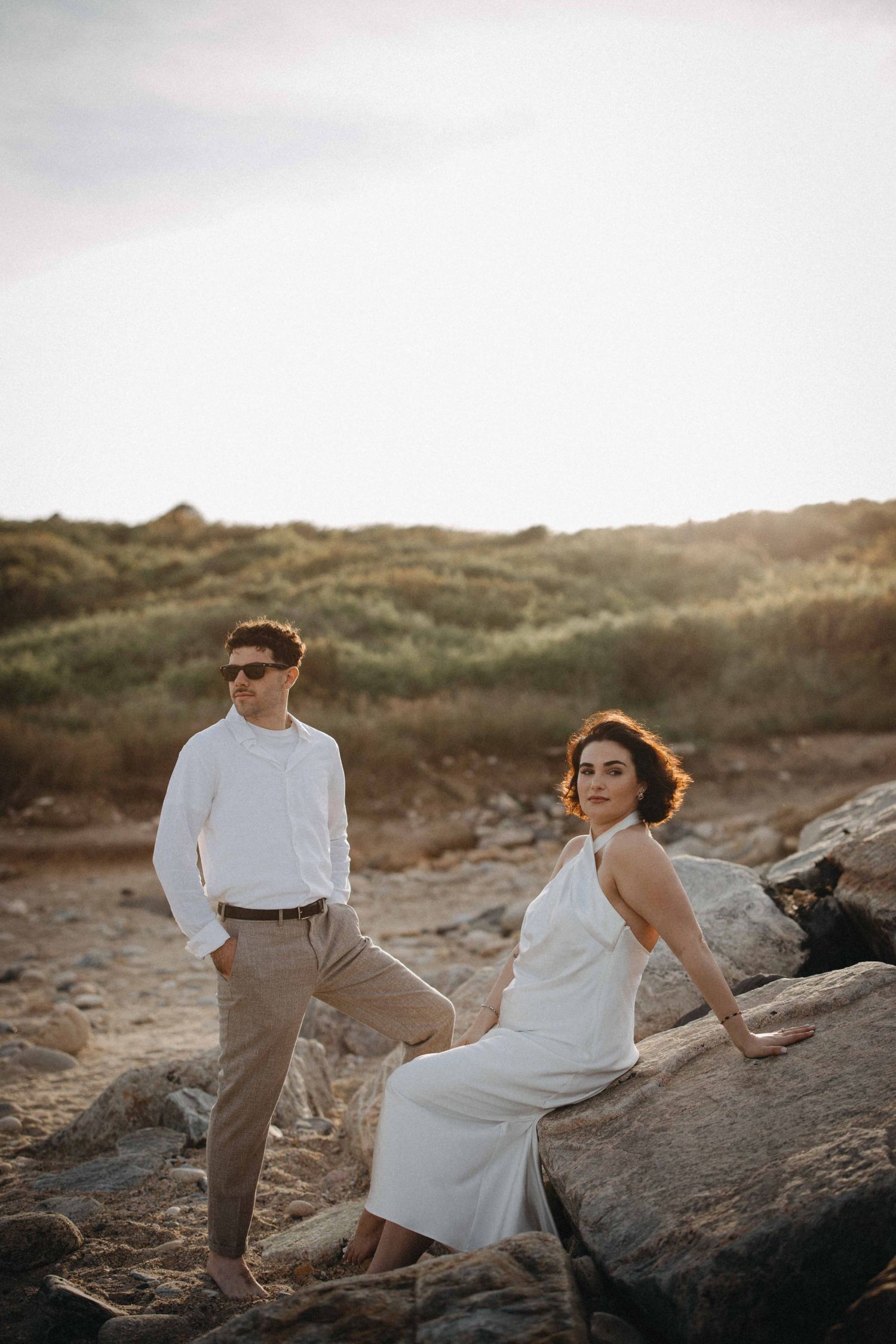 Couple posing on rocky beach at golden hour, woman in white dress seated on rocks, man in white shirt and khakis.
