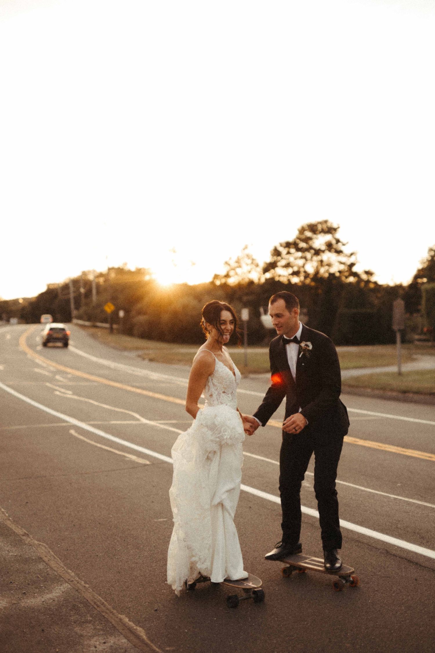 Bride and groom skateboarding together on a road at sunset on their wedding day.
