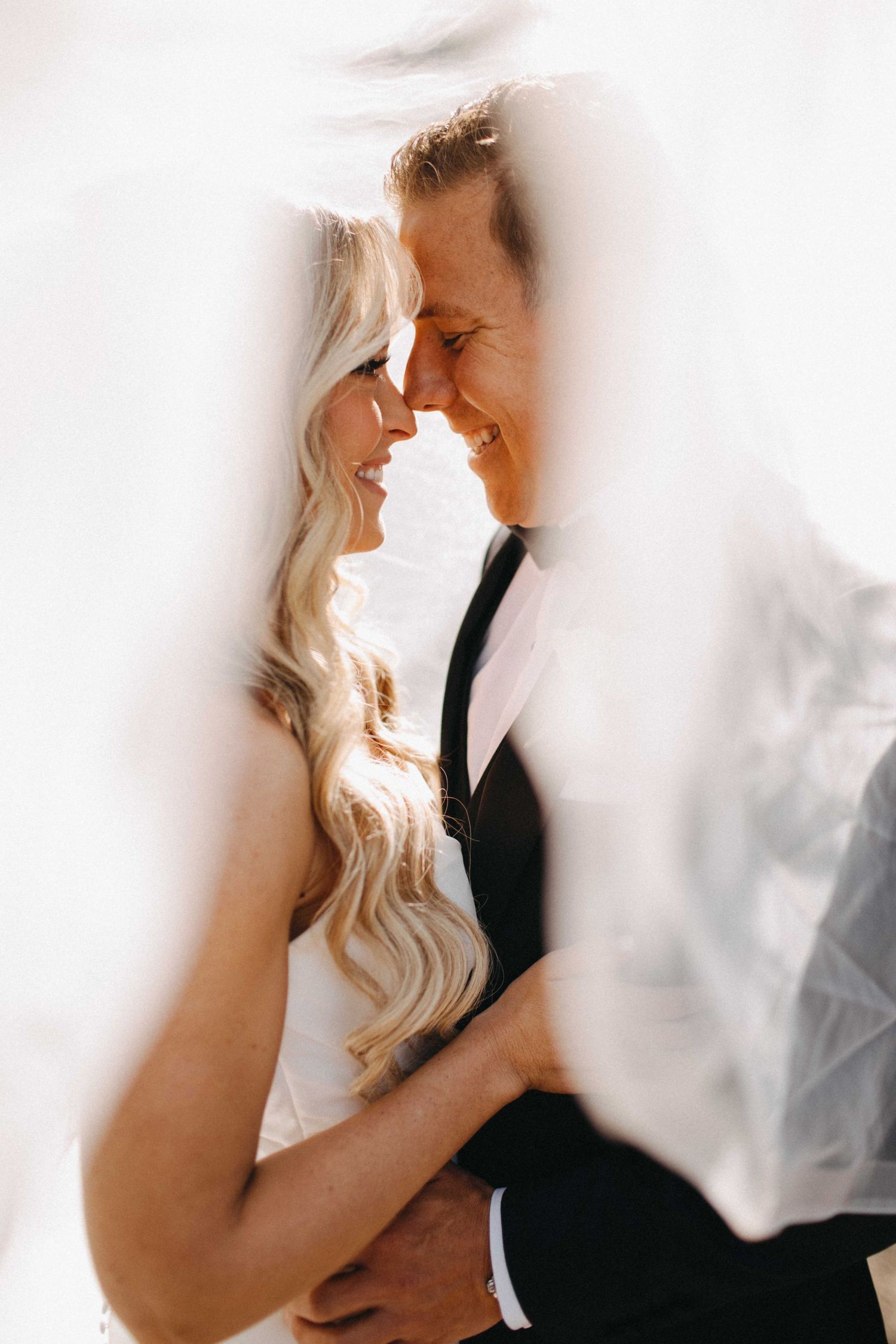 Bride and groom smiling forehead to forehead wrapped in flowing white veil in soft light.