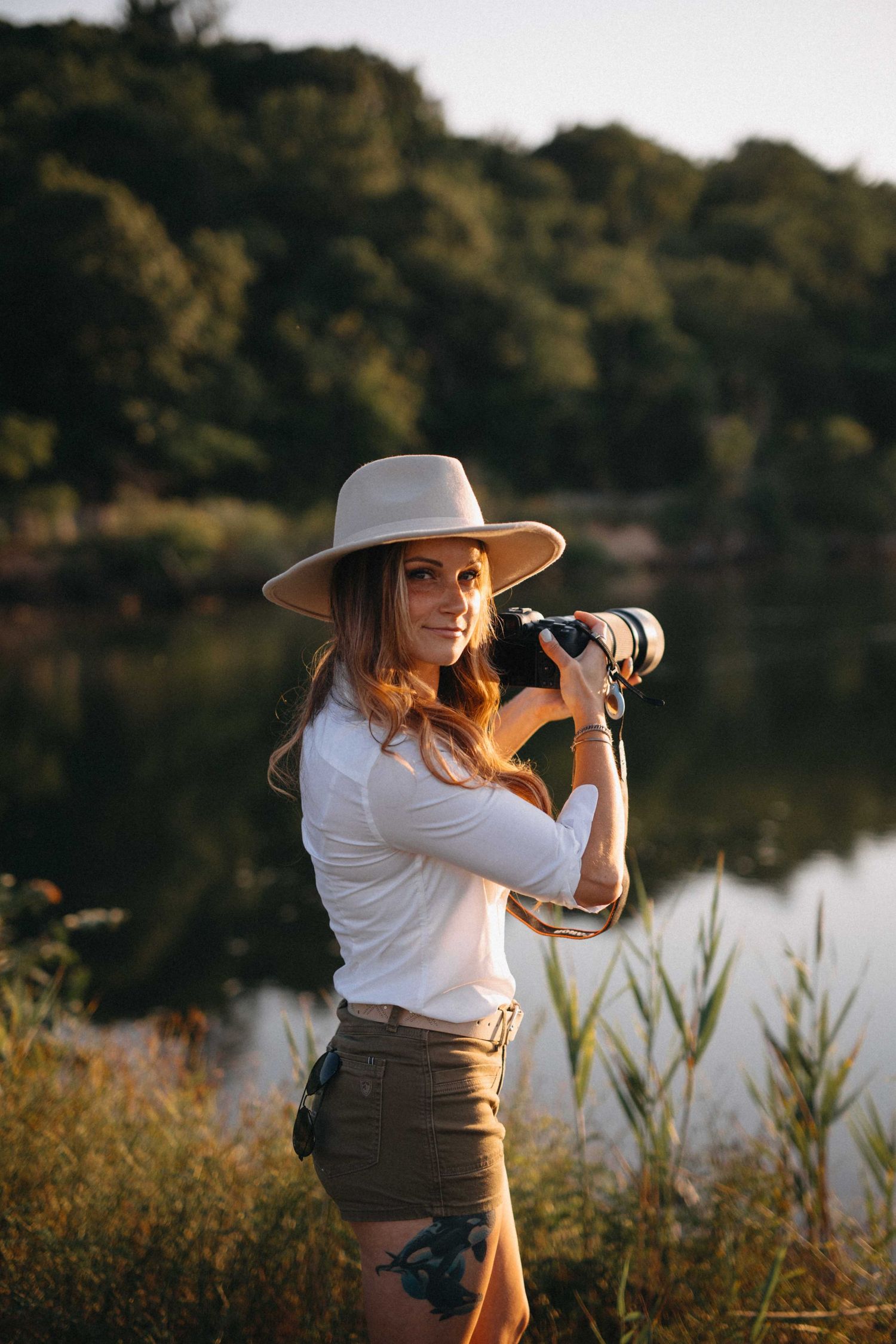 Woman in white shirt and hat holding a camera by a lake at golden hour in a natural outdoor setting.