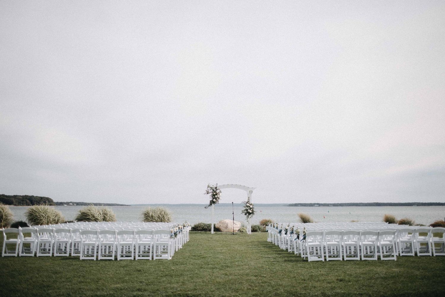 Outdoor waterfront wedding ceremony setup with white chairs and floral arch by the water.