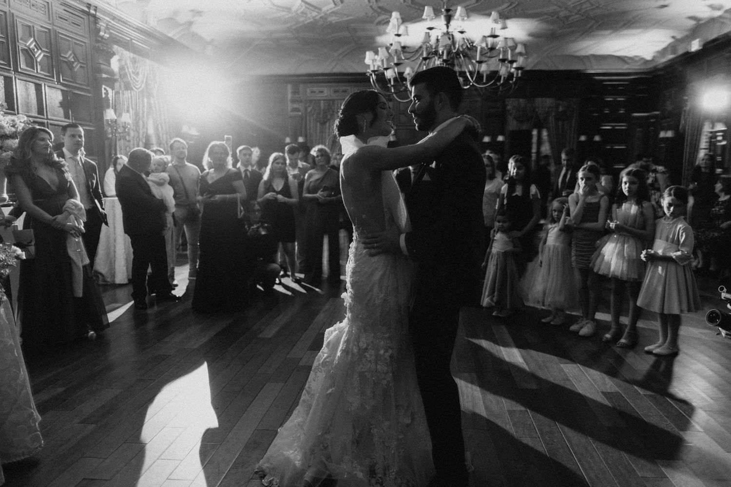 Black and white photo of bride and groom sharing their first dance surrounded by guests in an elegant ballroom.