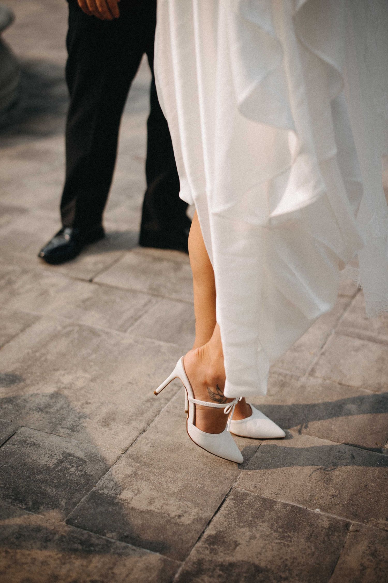 Bride wearing white pointed-toe heeled wedding shoes beside groom on stone pavement.