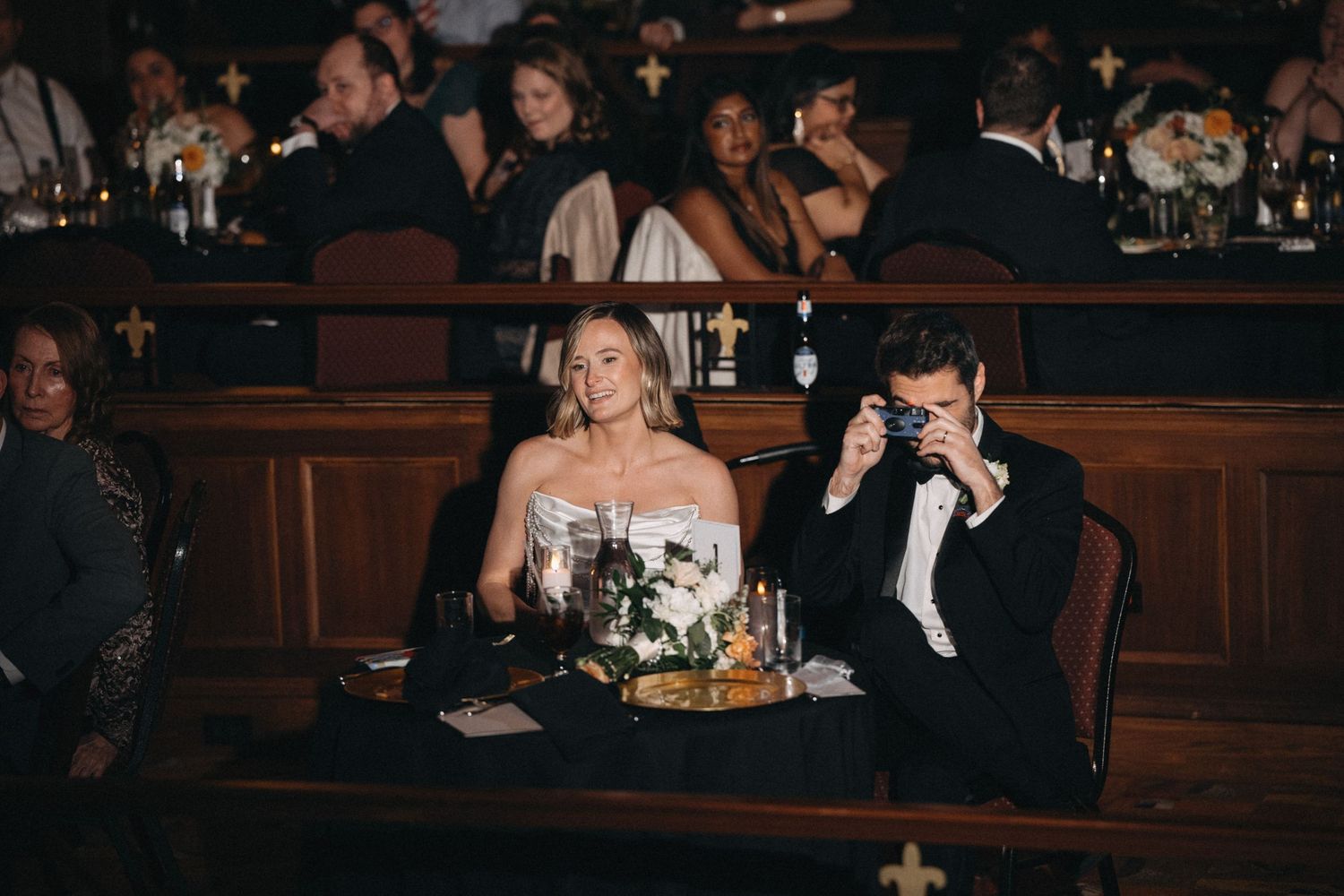 Bride and groom seated together at wedding reception in dark wood church pew venue.