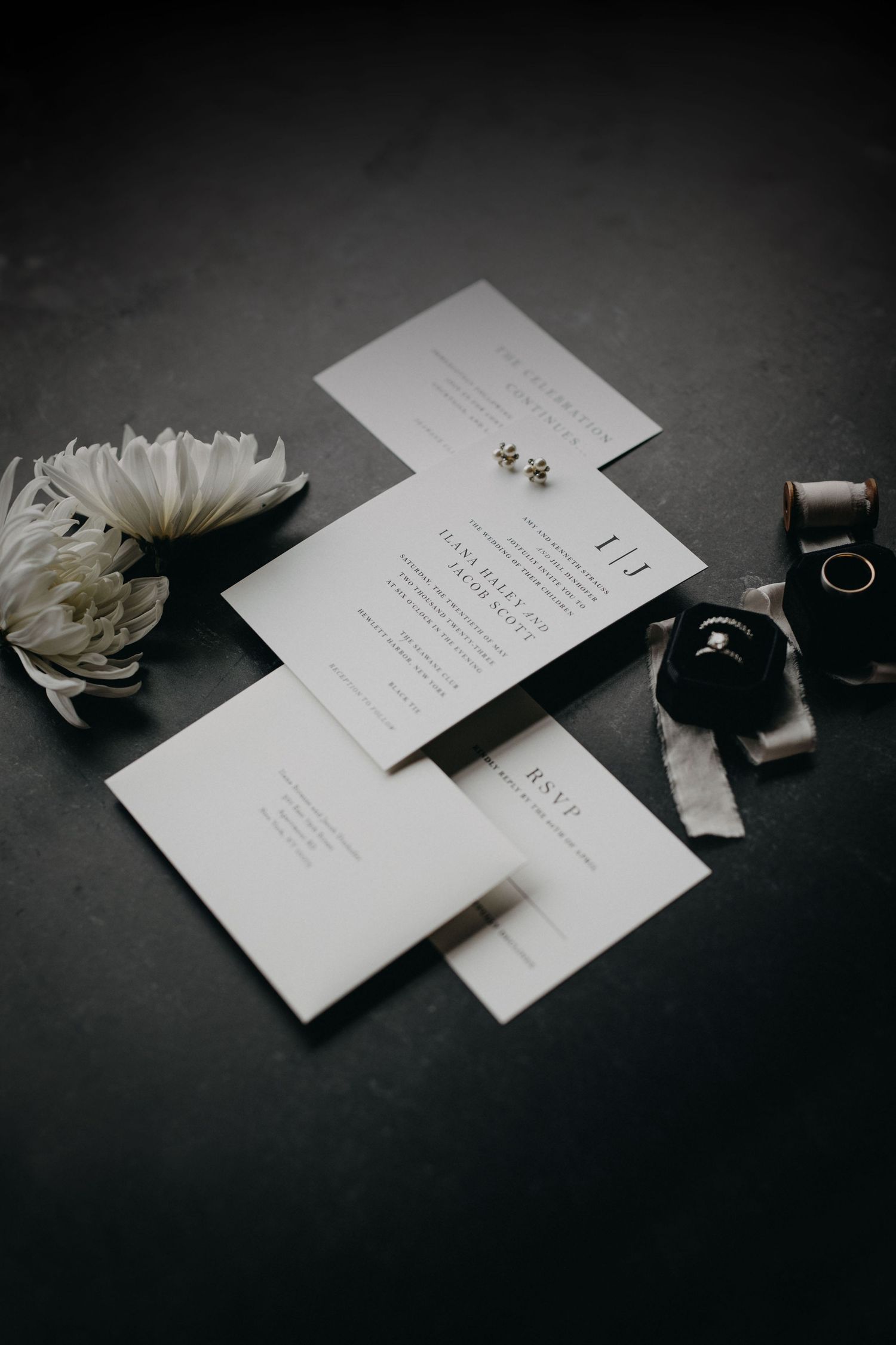 Elegant minimalist wedding stationery suite with white flowers, rings, and earrings on a dark background.