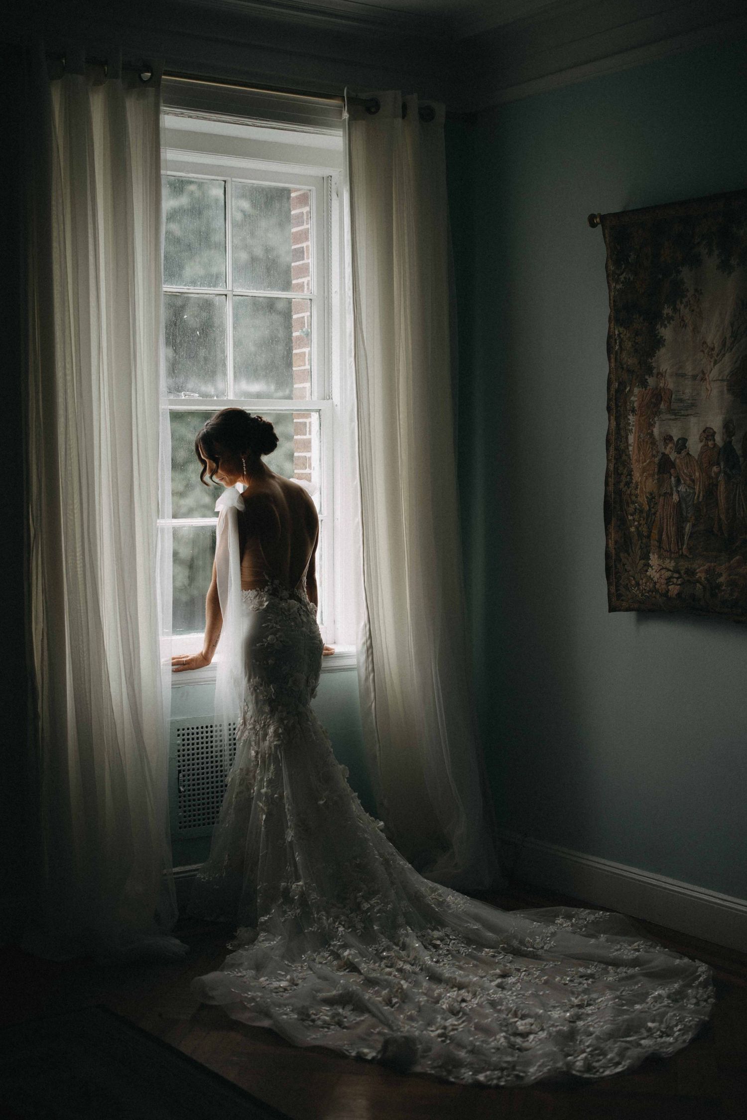 Bride in backless lace mermaid gown with long train stands silhouetted before a large window.