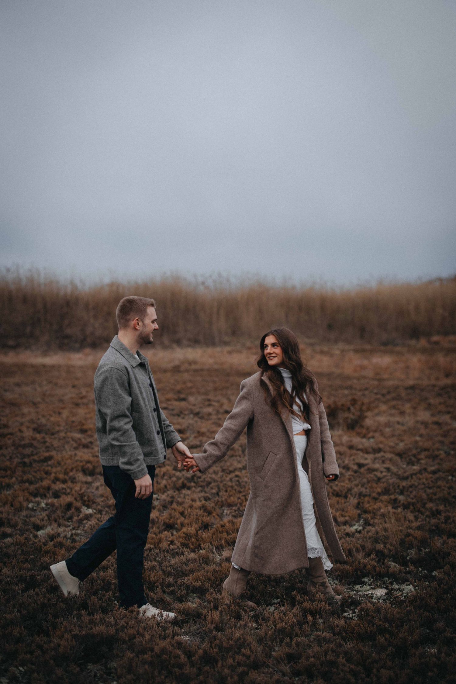 Couple holding hands and walking through a moody autumn field on an overcast day.