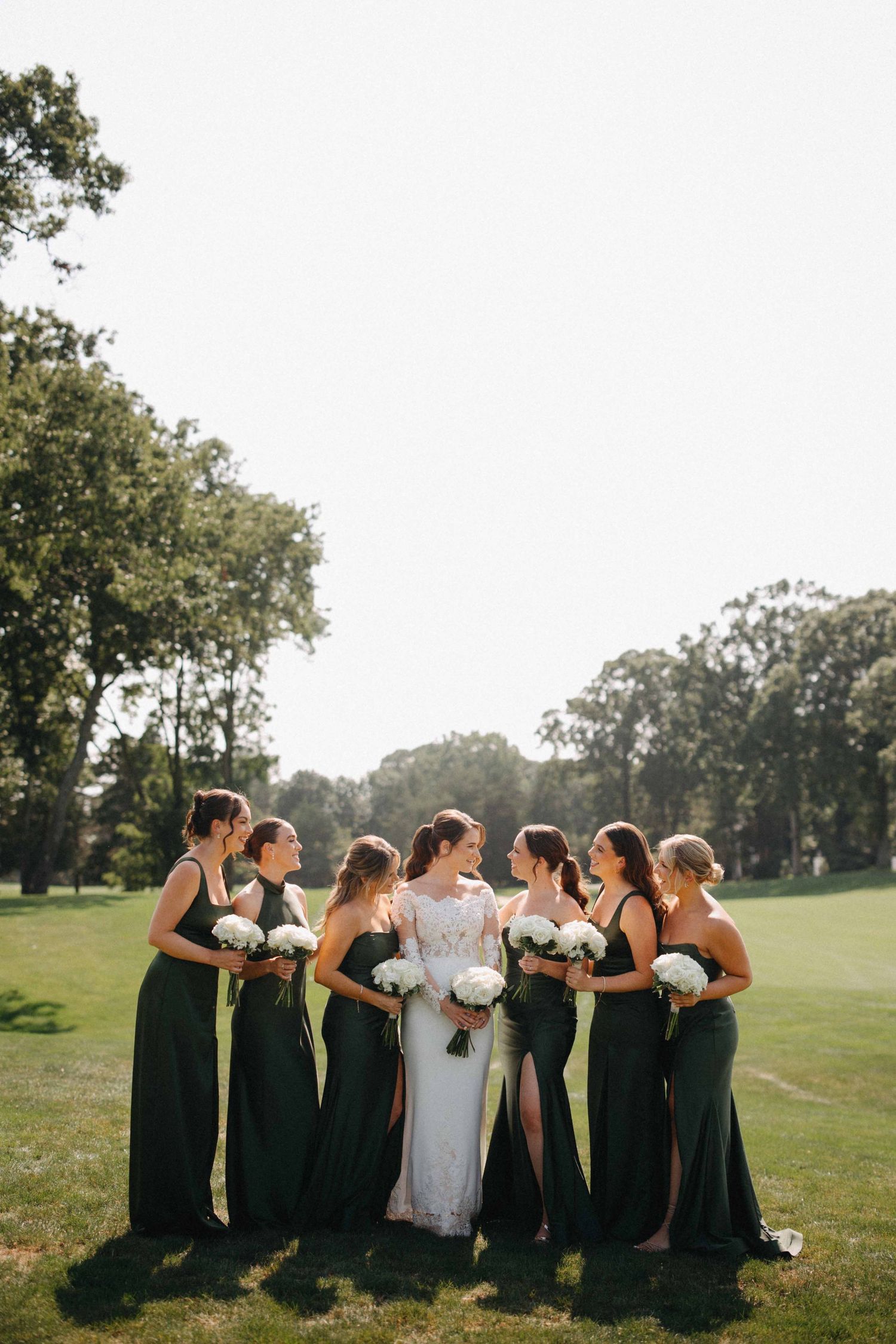 Bridesmaids in dark green gowns holding white bouquets surround lace-gowned bride on sunlit lawn.