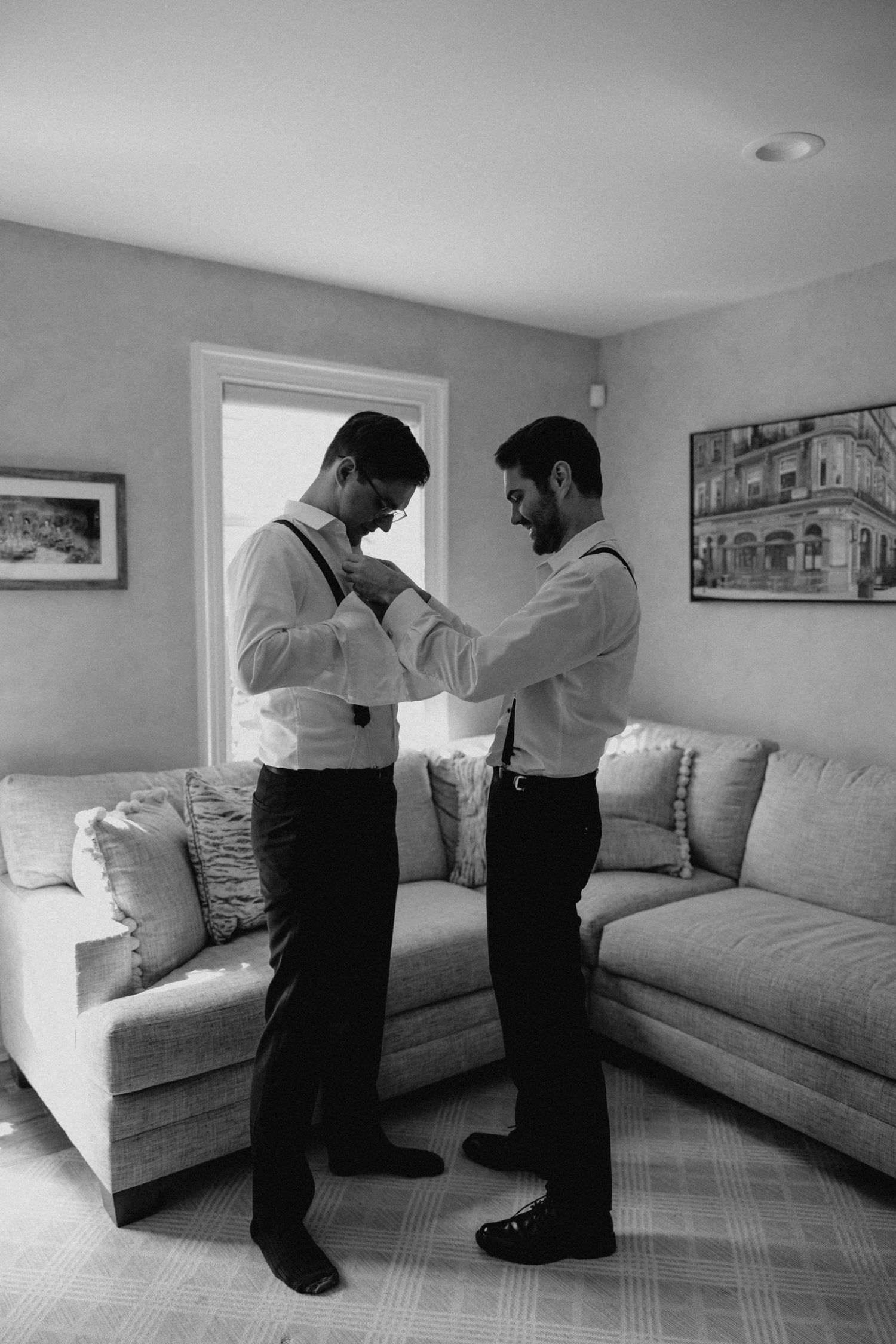 Two groomsmen help each other with ties and suspenders while getting ready in a bright living room.