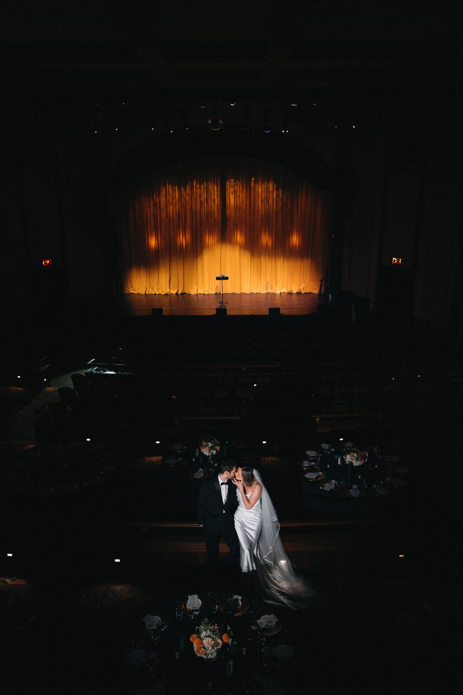 Bride and groom share a romantic moment before a glowing golden stage curtain in a dark theater.
