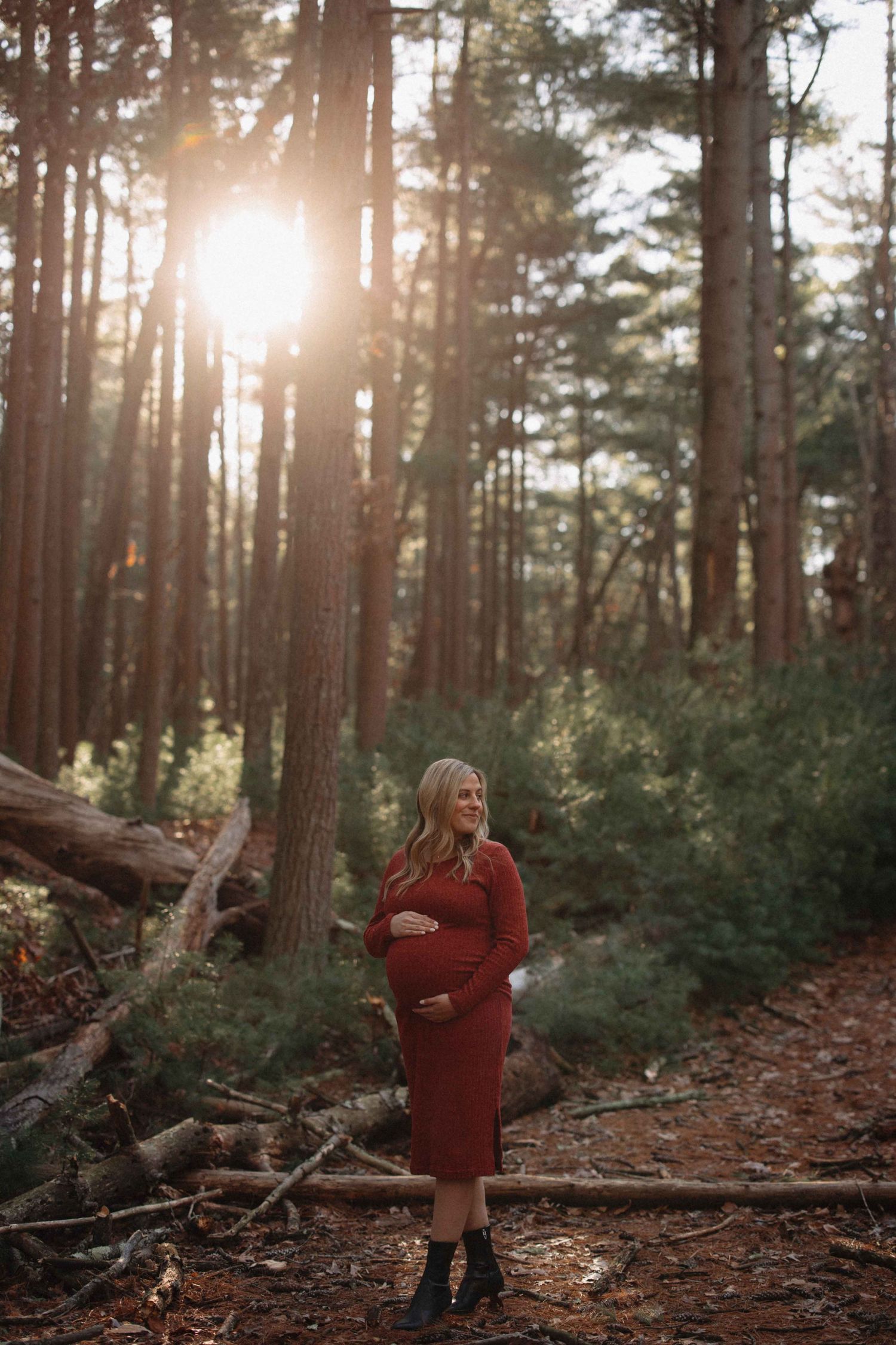 Pregnant woman in red dress cradling baby bump in sunlit forest during golden hour maternity photo session.