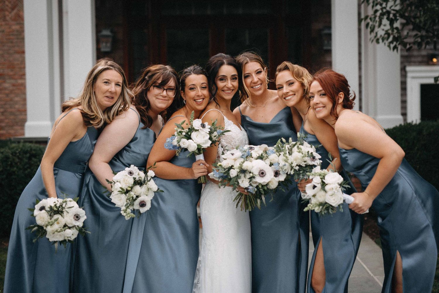 Smiling bride and six bridesmaids in steel blue gowns holding white floral bouquets posing together outdoors.