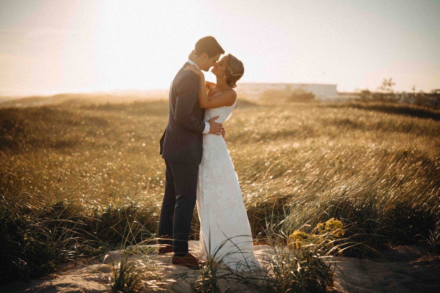 Bride and groom sharing a romantic kiss in golden sunset light among beach dunes.