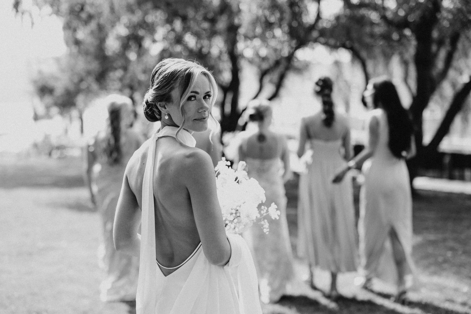 Black and white photo of bride in backless halter gown glancing back with bridesmaids behind her.