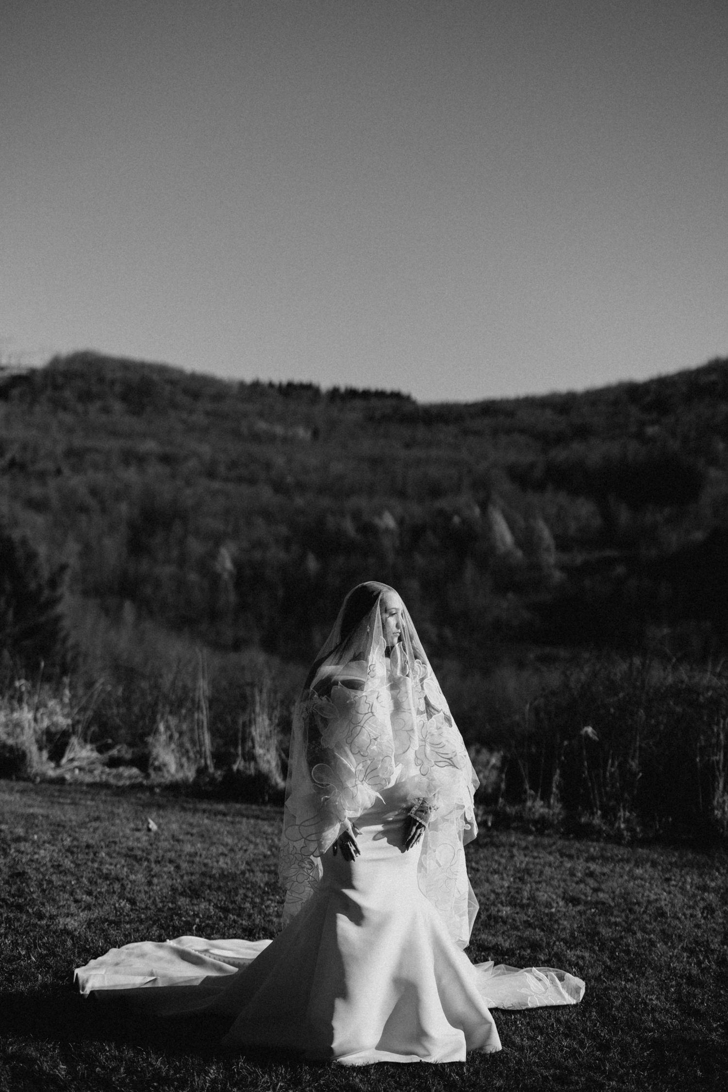 Black and white photo of a bride in a lace veil and gown posing in a scenic mountain landscape.