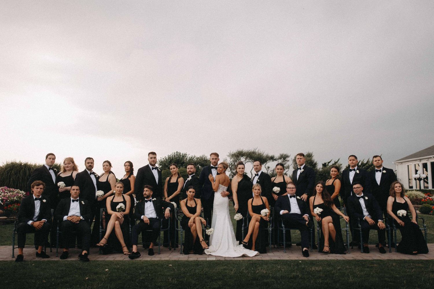 Large wedding party in black formal attire poses together on lawn under overcast sky.