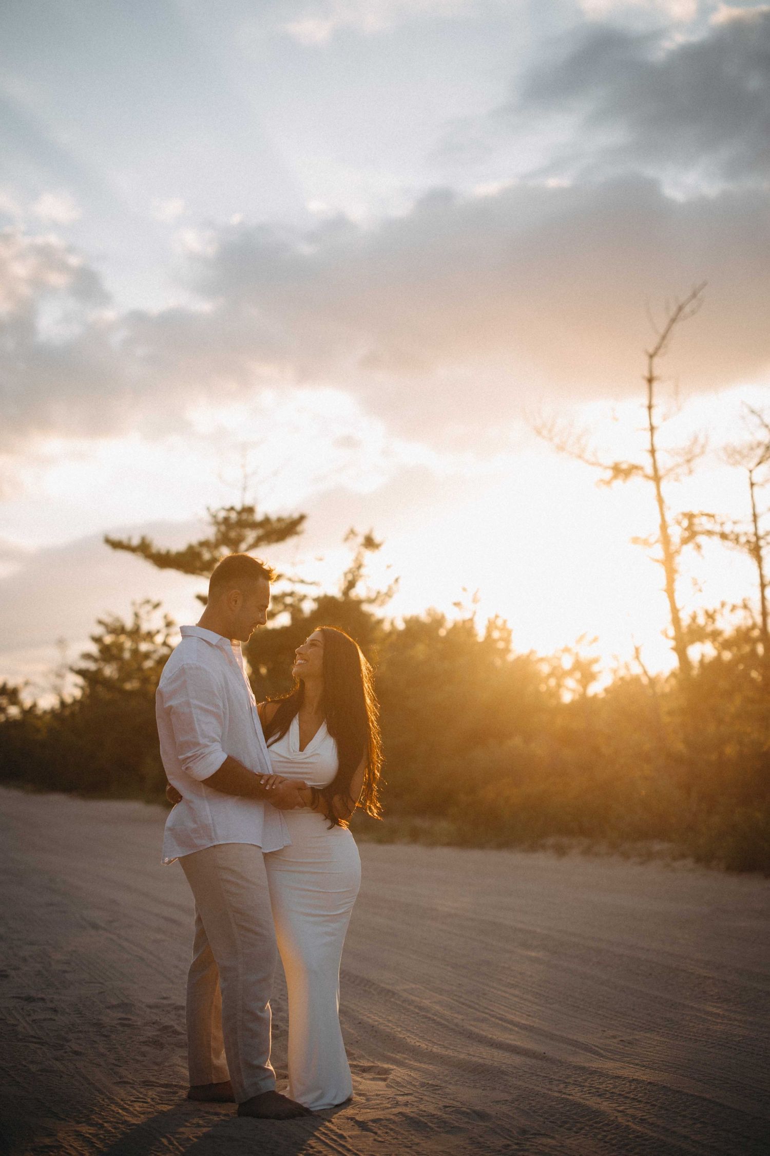 Romantic couple embracing on sandy beach with glowing golden sunset behind them.