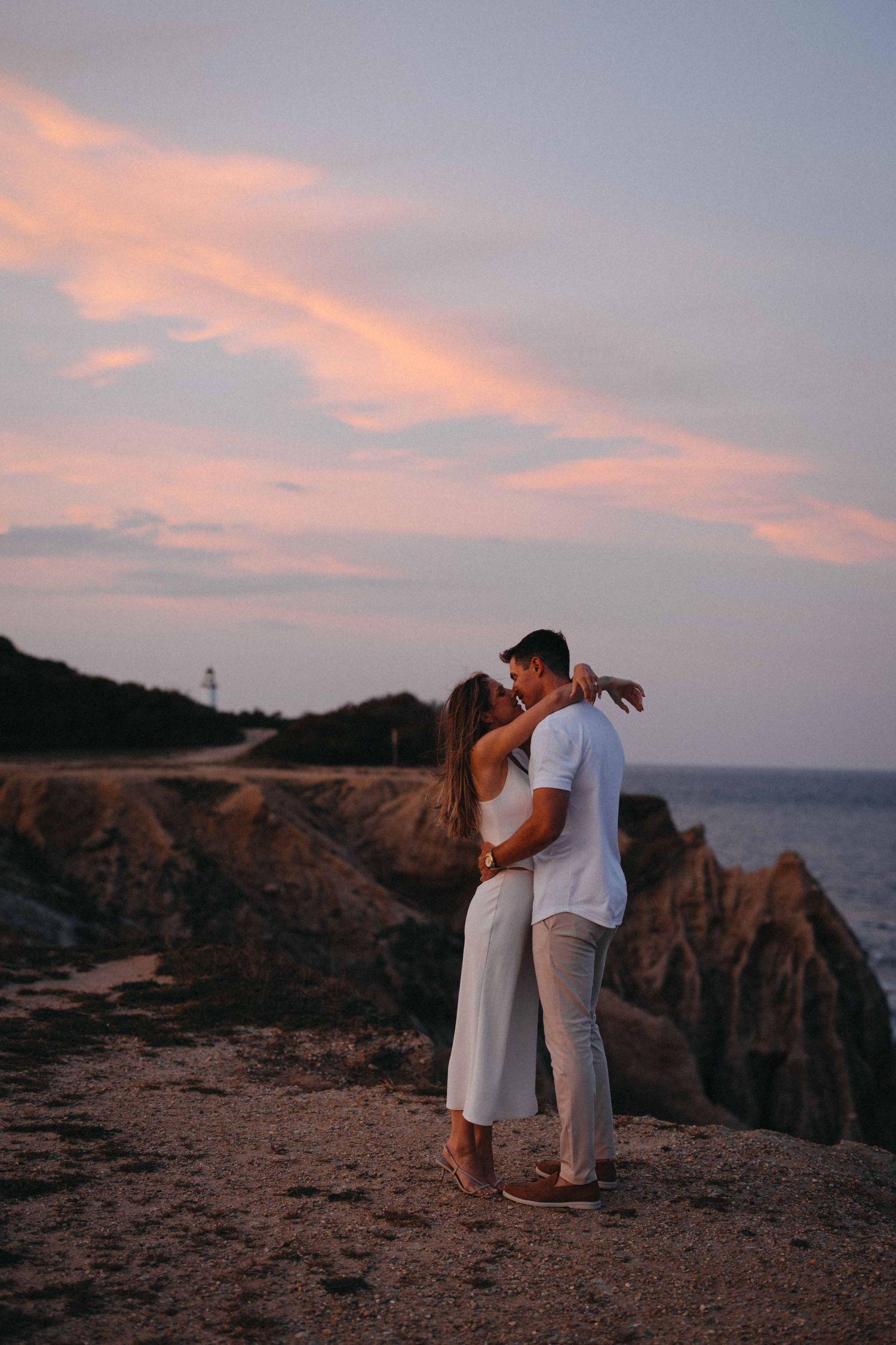 Couple sharing a romantic kiss on rocky coastal cliffs during a stunning pink and purple sunset.