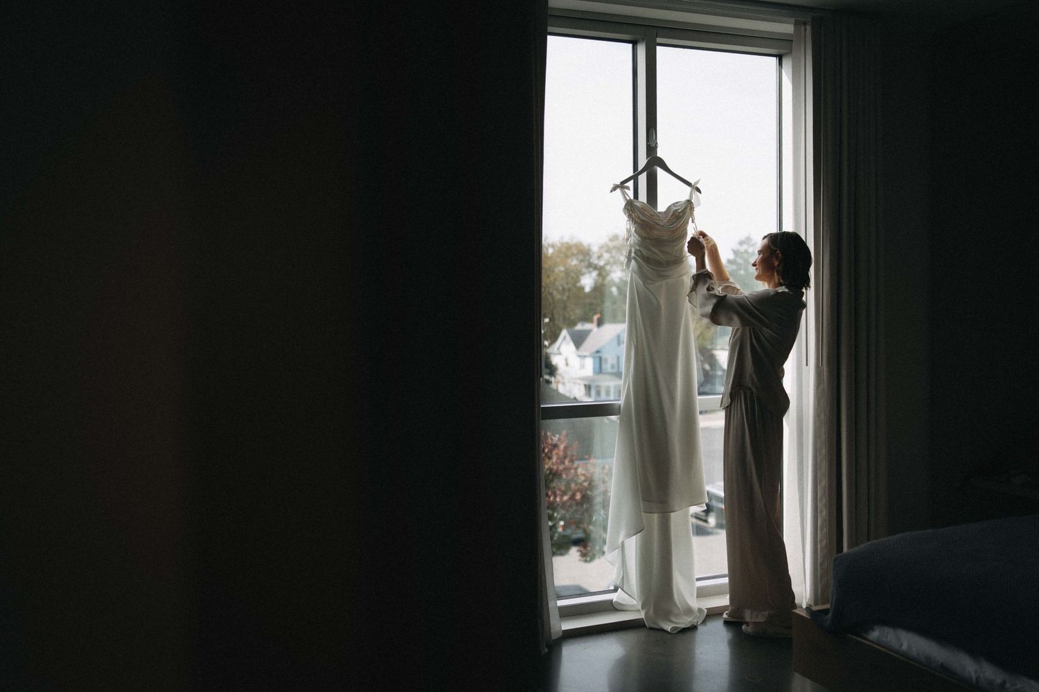 Bride in robe admiring her wedding dress hanging in a sunlit window before the ceremony.