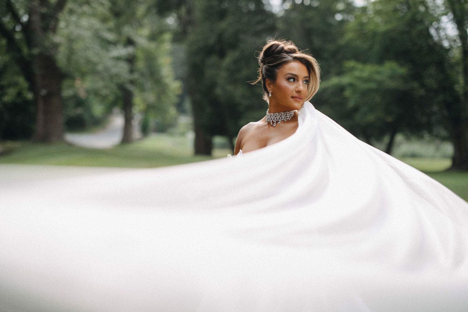 Bride in a strapless white gown with a dramatic flowing veil poses gracefully in a tree-lined outdoor setting.
