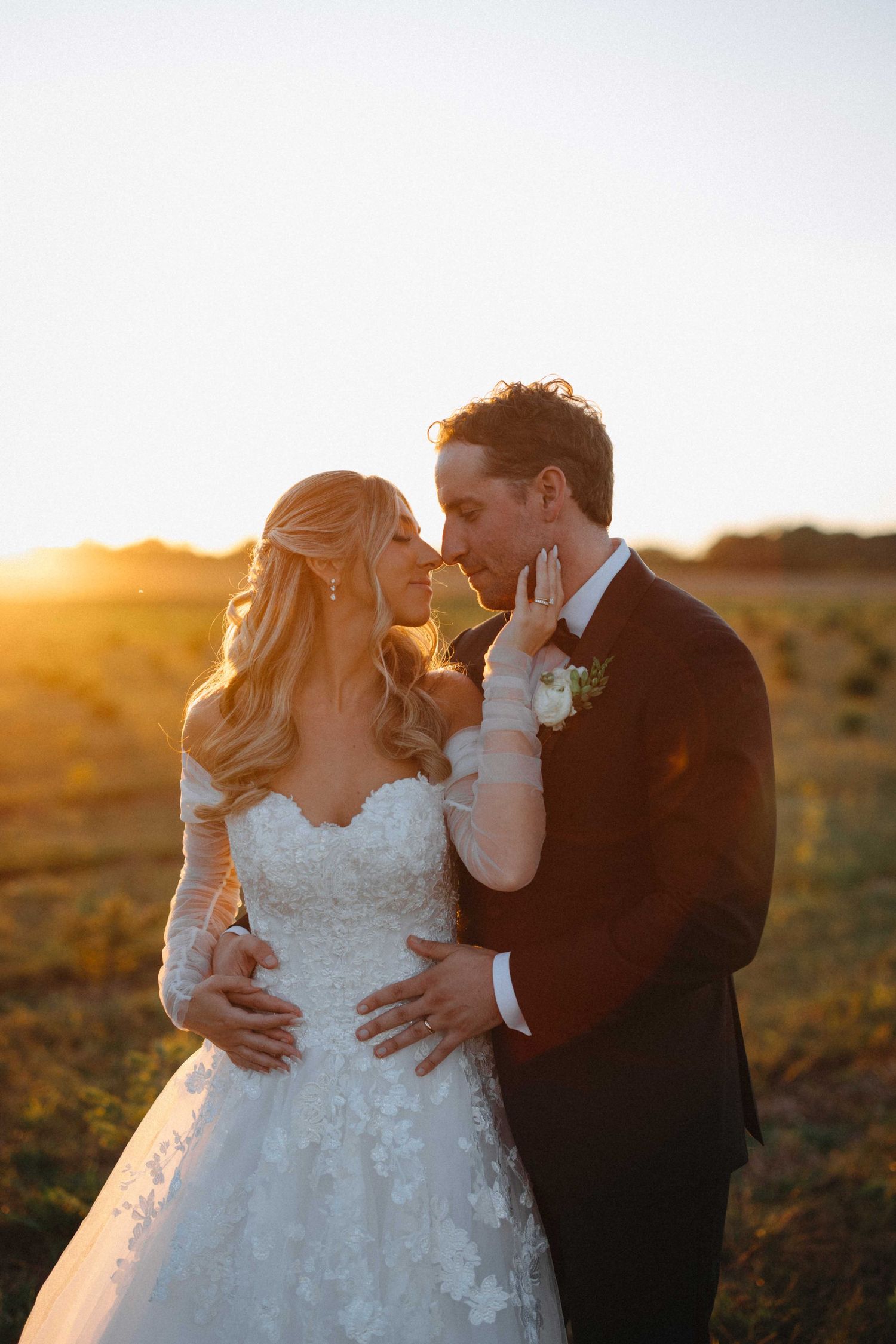 Bride and groom share romantic golden hour moment in open field, bride in lace off-shoulder gown.
