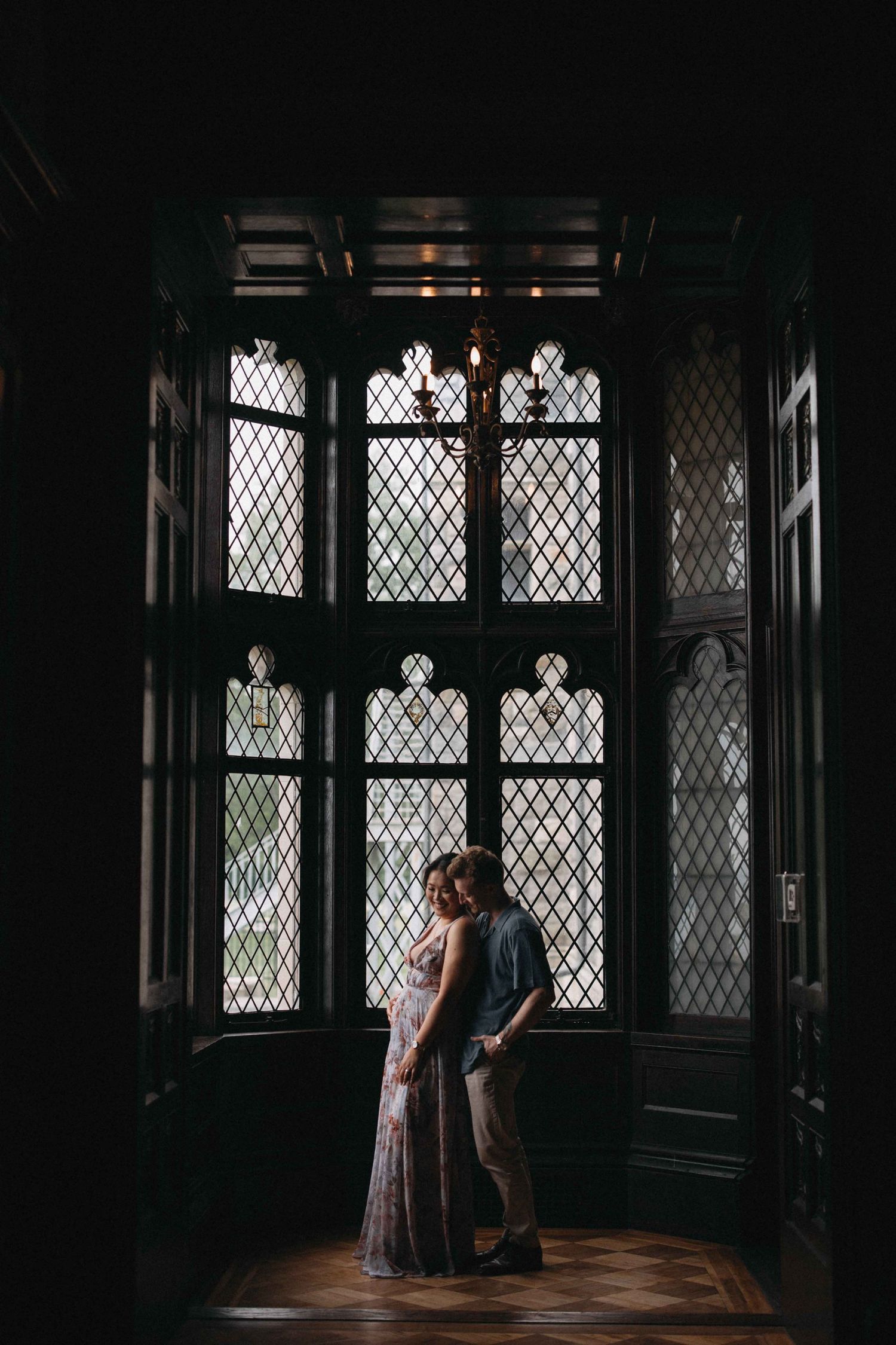 Couple standing close together in silhouette before dramatic gothic latticed windows in a dark interior.